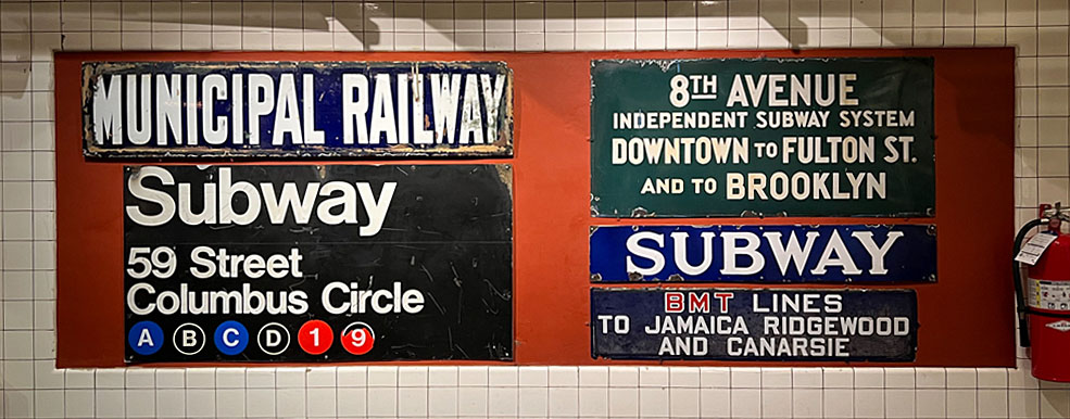 A collection of vintage subway signs, including "Municipal Railway," "Subway" for the 59th Street Columbus Circle station, and signs for the 8th Avenue Independent Subway System. The signs feature bold fonts and iconic colors, such as black, white, and green.