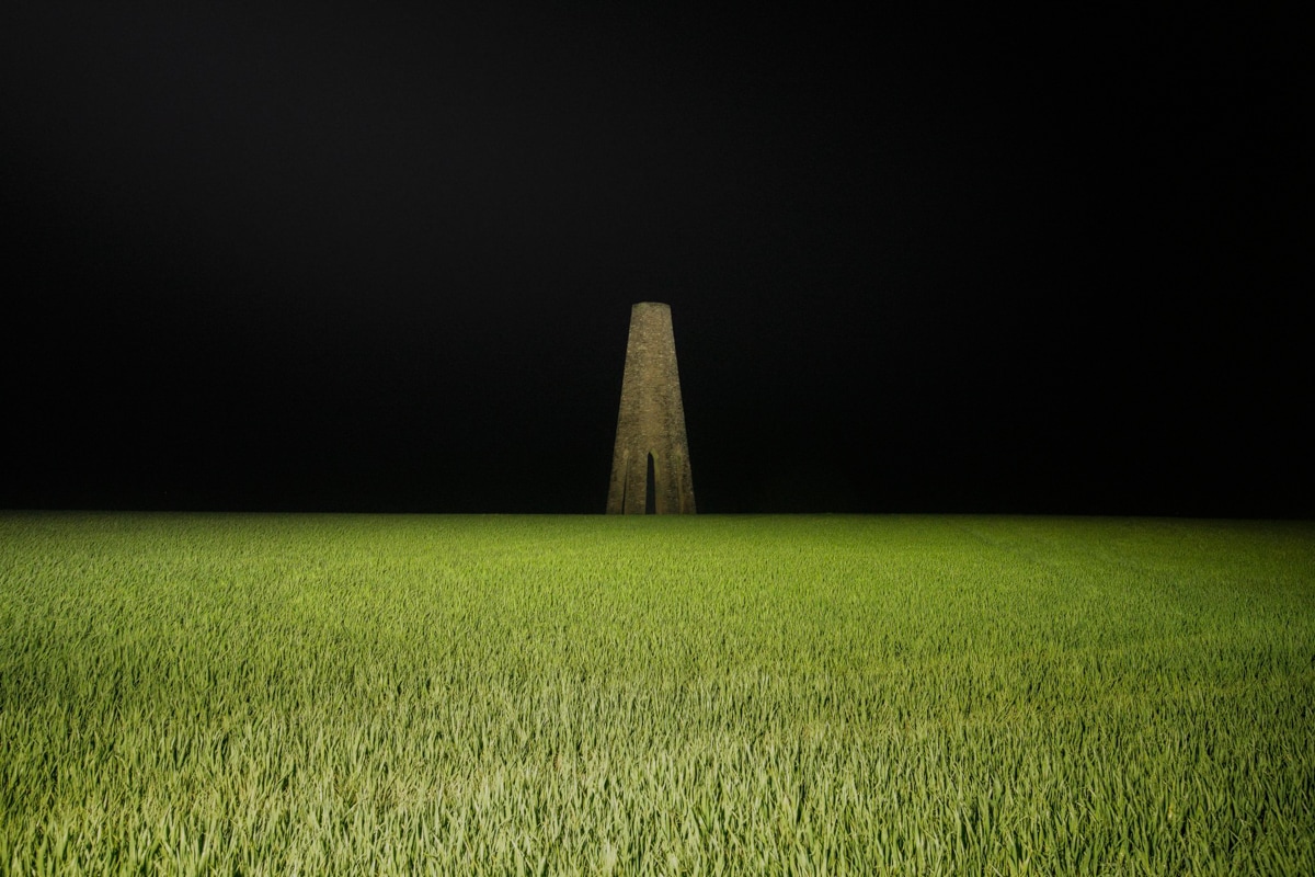 A surreal night photograph showing a tall, narrow stone structure rising from a vast green field, illuminated by soft lighting. The stark contrast between the brightly lit grass and the pitch-black sky creates an eerie, otherworldly atmosphere, with the solitary tower-like structure as the central focus.