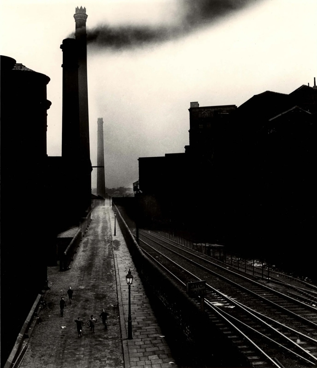 A black-and-white photograph of an industrial street scene, likely from mid-20th century Britain. A group of children walk along a cobbled road flanked by looming factory buildings. Two tall smokestacks dominate the skyline, with one releasing a thick plume of black smoke. Railway tracks run parallel to the road on the right side, bordered by a stone wall. The image is atmospheric, with heavy contrast between the smoke-darkened sky and the silhouettes of industrial structures.