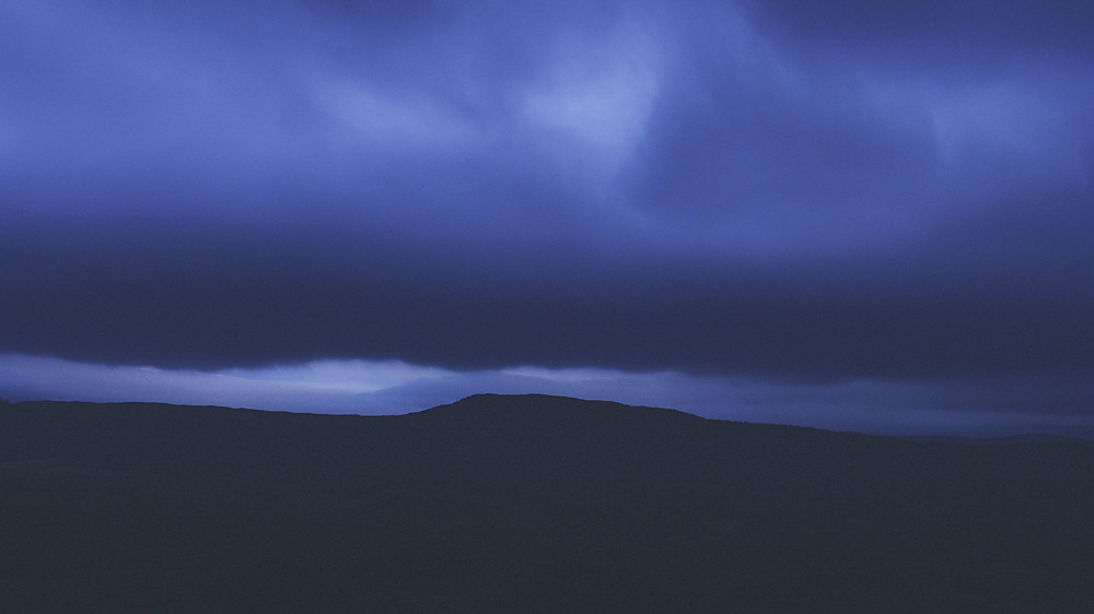Clouds cascade over a dark mountain ridge in a dramatic display, creating the appearance of misty waterfalls beneath a pale blue sky.