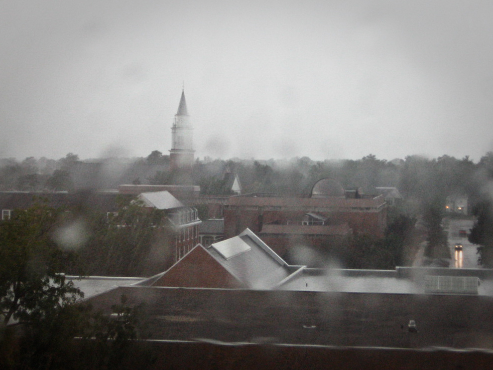 This photograph shows a rainy, overcast day with a view of a town or campus. The scene is blurred by raindrops on the window, adding to the dreary and misty atmosphere. In the background, a tall spire is visible, partially obscured by the rain and fog, with rooftops and trees in the foreground. The wet streets reflect the dim light of the day, and the overall mood feels calm yet somber, capturing the essence of a stormy or rainy afternoon. The muted colors and softened outlines enhance the feeling of rain-soaked tranquility.