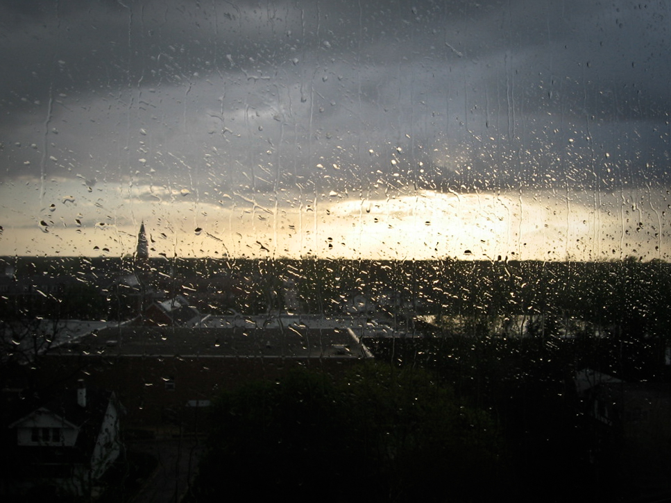 This photograph captures a rainy day, with raindrops covering a window, through which the distant landscape is visible. The sky is overcast, and the sun is low on the horizon, casting a faint glow and illuminating the rain-splattered window. A silhouette of a tall spire can be seen in the background, surrounded by rooftops and trees. The contrast between the gloomy weather and the soft light breaking through the clouds adds a melancholic yet peaceful feeling to the scene. The focus on the raindrops creates a textured and immersive effect.