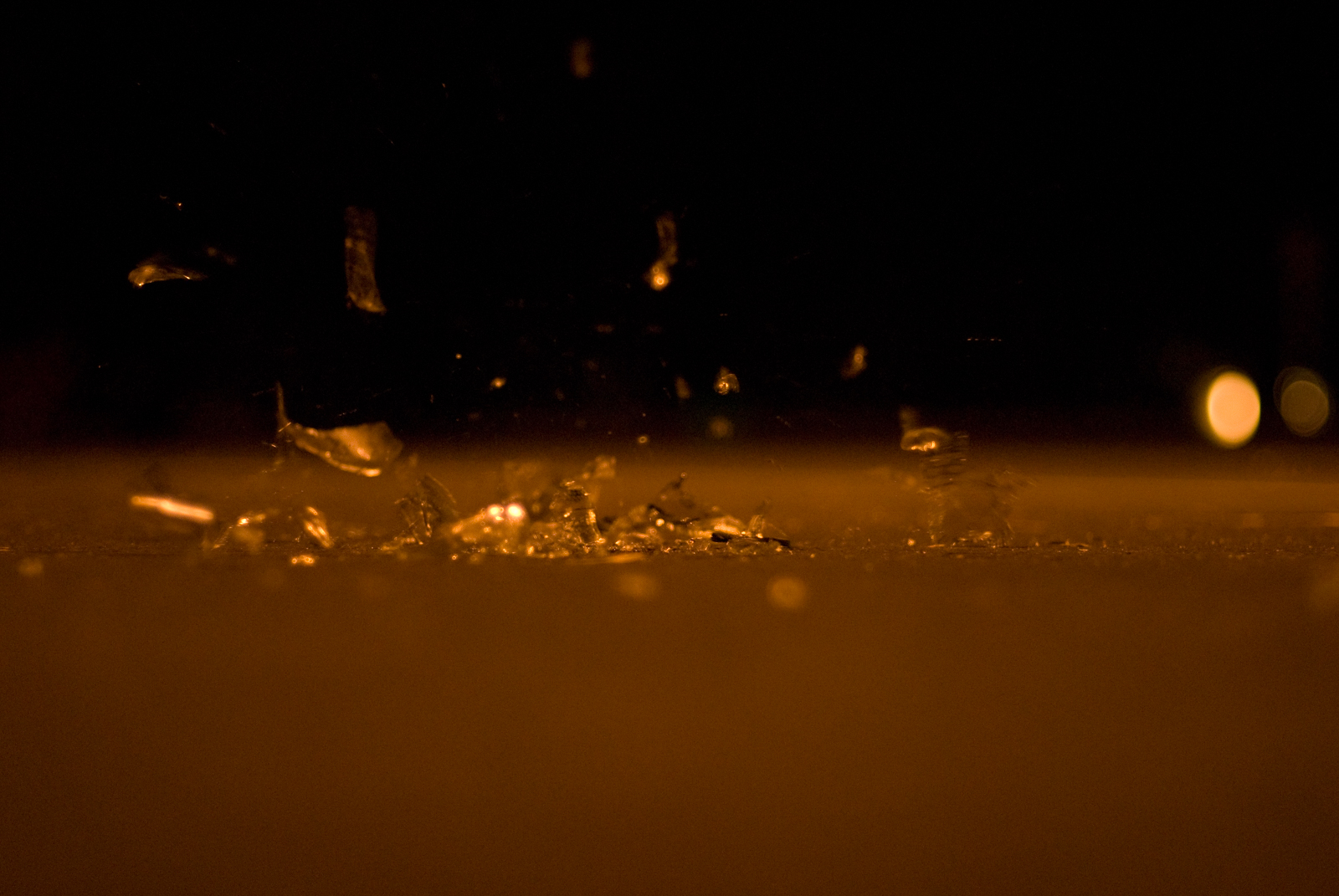 This close-up photograph captures shattered glass pieces scattered across the ground, with a few fragments suspended in mid-air, as if just recently broken. The scene is bathed in warm, golden lighting, creating a striking contrast between the sharp glass shards and the smooth, blurred background. The faint bokeh lights in the distance add a sense of depth and atmosphere. The image conveys a moment of action and fragility, emphasizing the delicate nature of the glass and the quiet stillness after a sudden break.