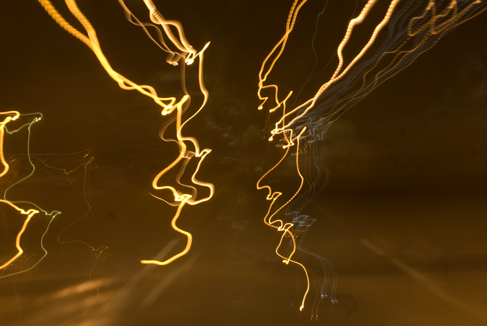 This abstract photograph showcases jagged, swirling streaks of yellow, orange, and white light against a dark background. The light trails seem to twist and dance across the frame, forming chaotic, erratic patterns that suggest movement and energy. The overall effect is dynamic and mesmerizing, with the flowing, wave-like lines creating a sense of spontaneity and unpredictability. The dark backdrop contrasts sharply with the glowing light, making the streaks stand out prominently, while their irregular shapes add to the abstract, dreamlike quality of the image.