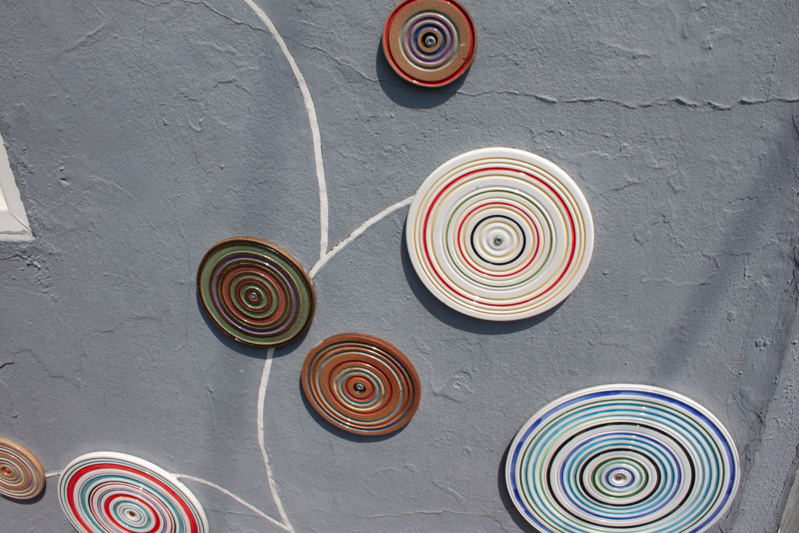 This image features a decorative installation of circular ceramic plates or discs mounted on a gray textured wall. Each disc has concentric rings in various colors, such as red, green, blue, and brown, creating a visually striking pattern. The design on the wall resembles an abstract tree, with white lines connecting the discs, mimicking branches or stems. The vibrant colors and geometric shapes stand out against the muted gray background, giving the installation a modern, artistic feel. The overall effect is playful and creative, adding an element of visual interest to the otherwise plain wall.