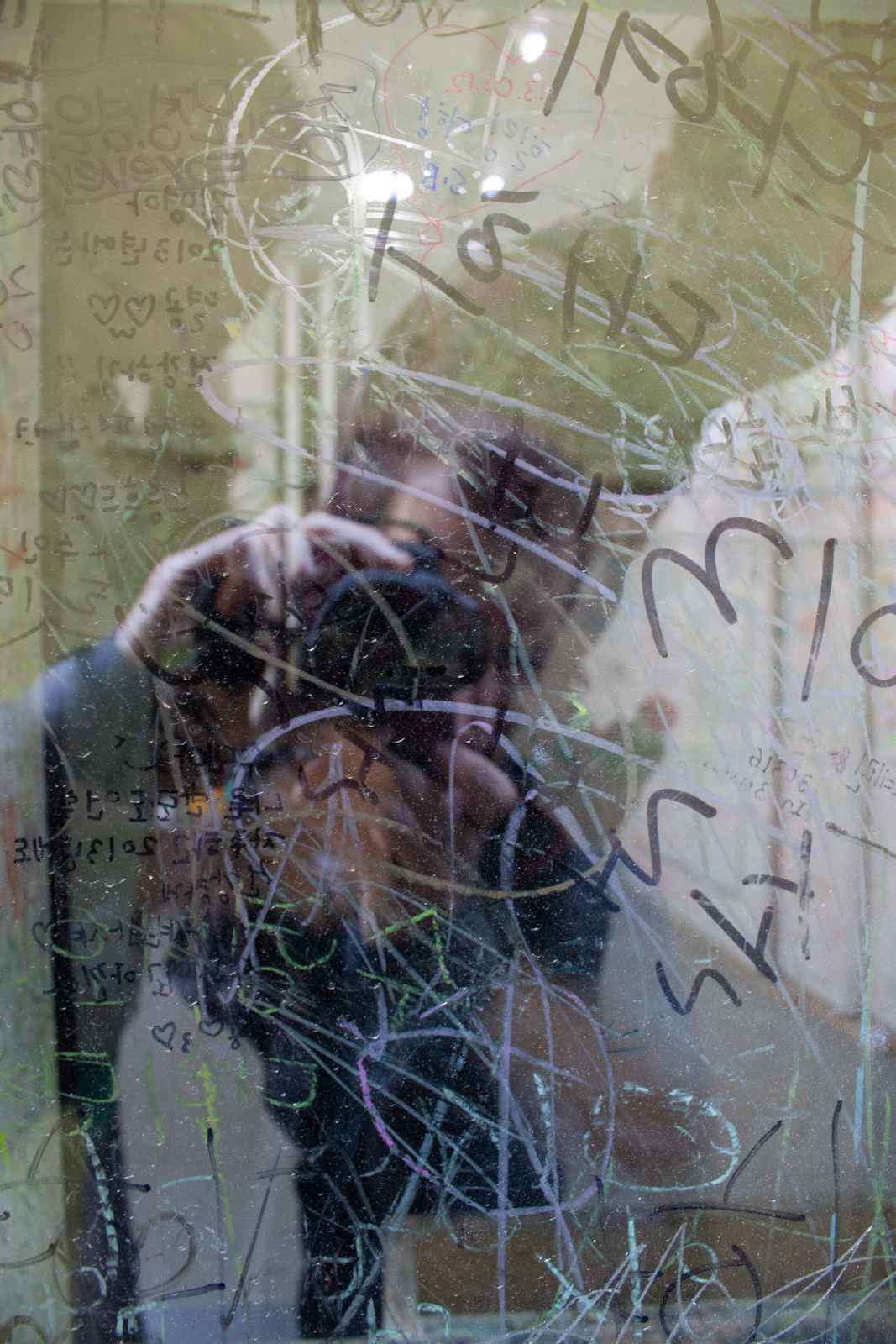 This image features a reflection of the photographer in a scratched and graffiti-covered glass surface. The glass is marked with various scribbles, drawings, and writings, creating an abstract and chaotic pattern. The layered effect of the reflections and the markings gives the image a sense of depth and mystery, as the viewer can partially make out the photographer’s form behind the glass. The interaction between the viewer and the artwork through the reflection suggests themes of identity, perception, and interaction with urban spaces, where public markings become part of the visual landscape. The overall effect is a mix of personal and impersonal, as the reflective surface both reveals and obscures at the same time.