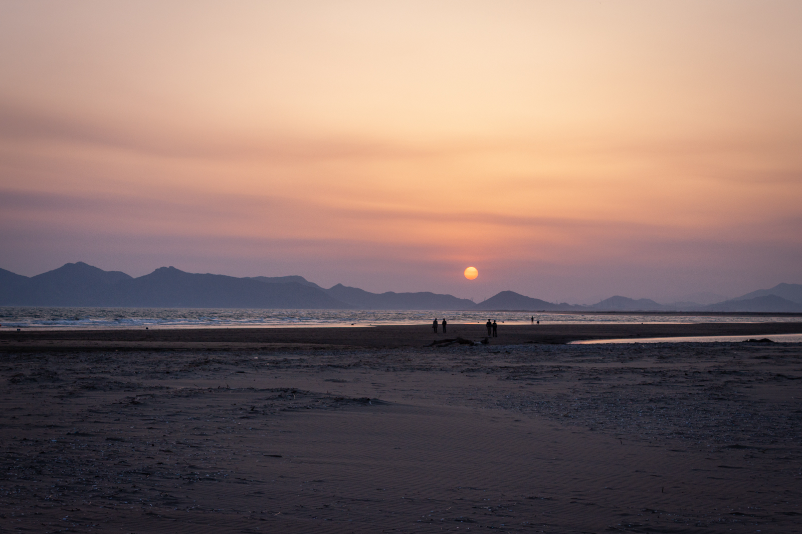 This image captures a serene sunset over a beach, with the sun hanging low in the sky as it casts a warm orange and purple glow across the horizon. The distant mountains are silhouetted against the soft gradient of colors in the sky, adding depth to the scene. The calm sea reflects the fading sunlight, creating a tranquil atmosphere. In the foreground, a few people can be seen walking along the beach, their small figures emphasizing the vastness of the landscape. The gentle waves and the expansive sand add to the peacefulness of the scene, evoking a sense of quiet reflection and beauty in nature.
