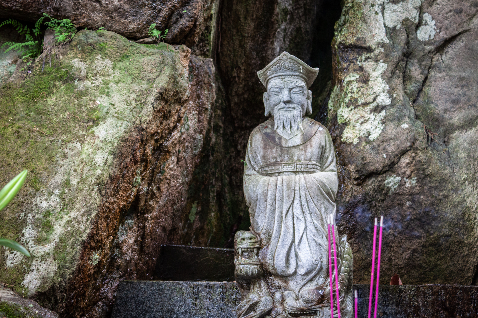A small stone statue of a bearded man with a traditional hat, placed in front of a rocky backdrop, surrounded by moss and incense sticks burning in front of it.