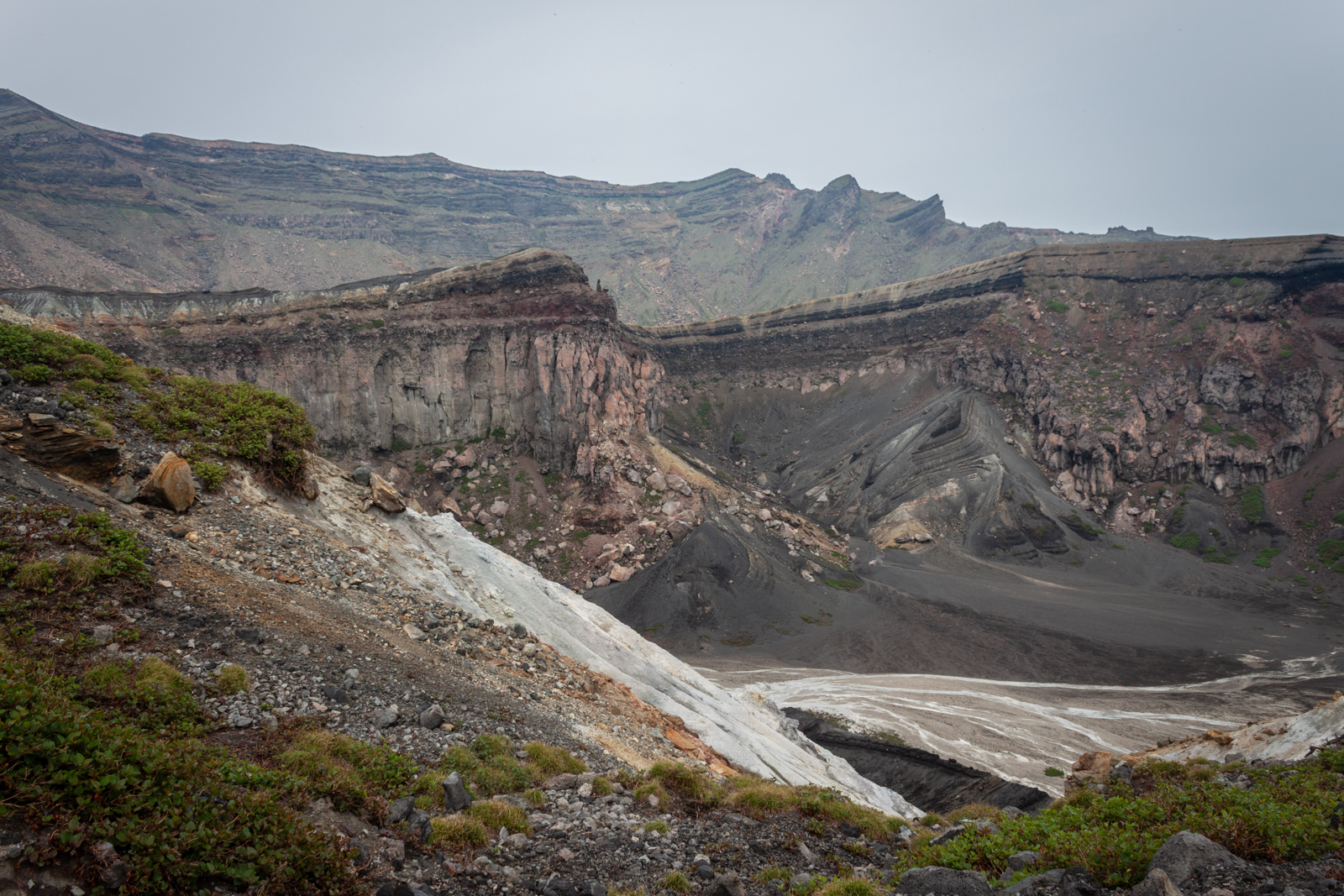 A view of rugged, steep cliffs and ash-laden slopes of a volcanic crater, showcasing layers of rock formations and patches of greenery.