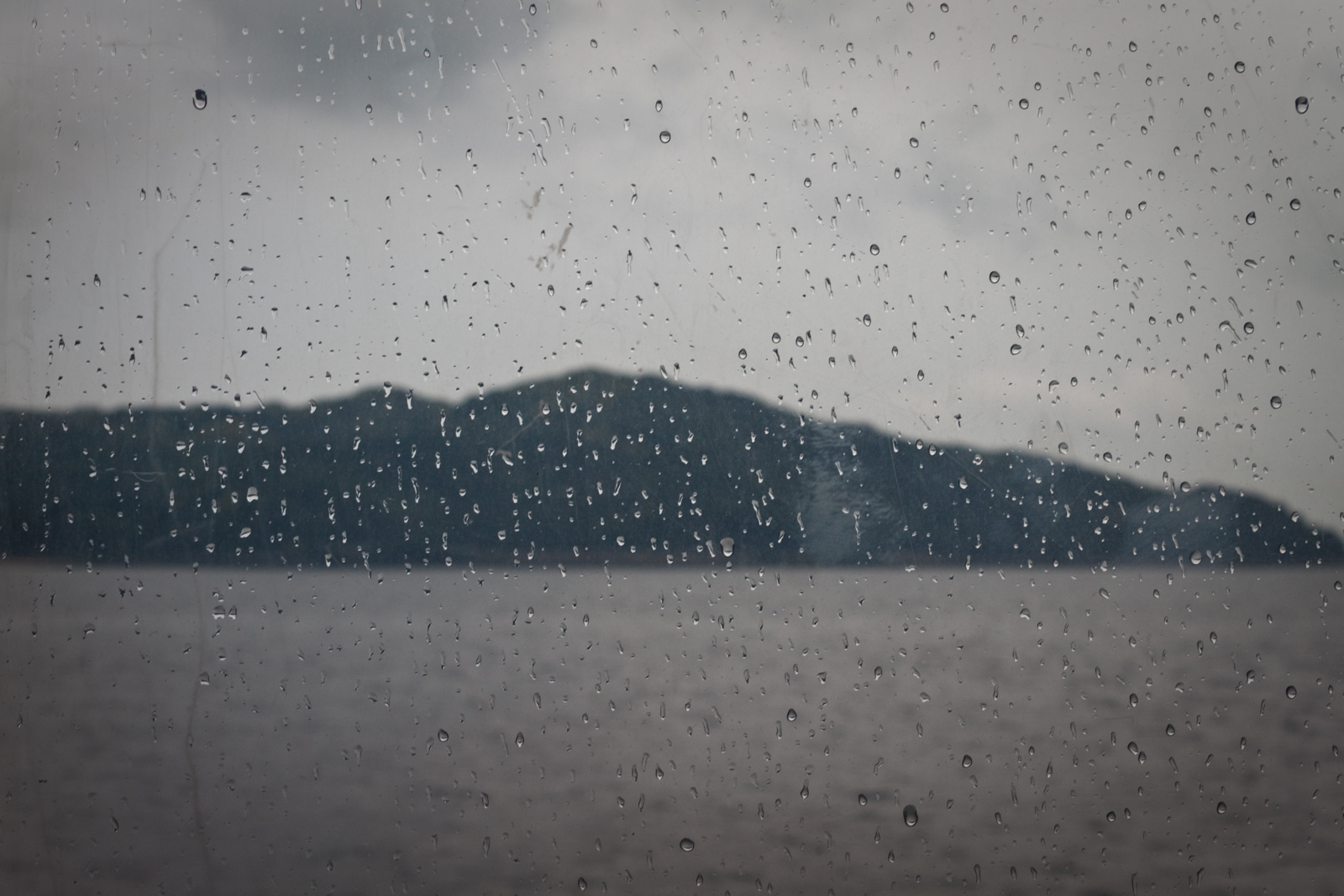 Raindrops cover a window, blurring the view of a distant island across a body of water, under a cloudy, overcast sky.