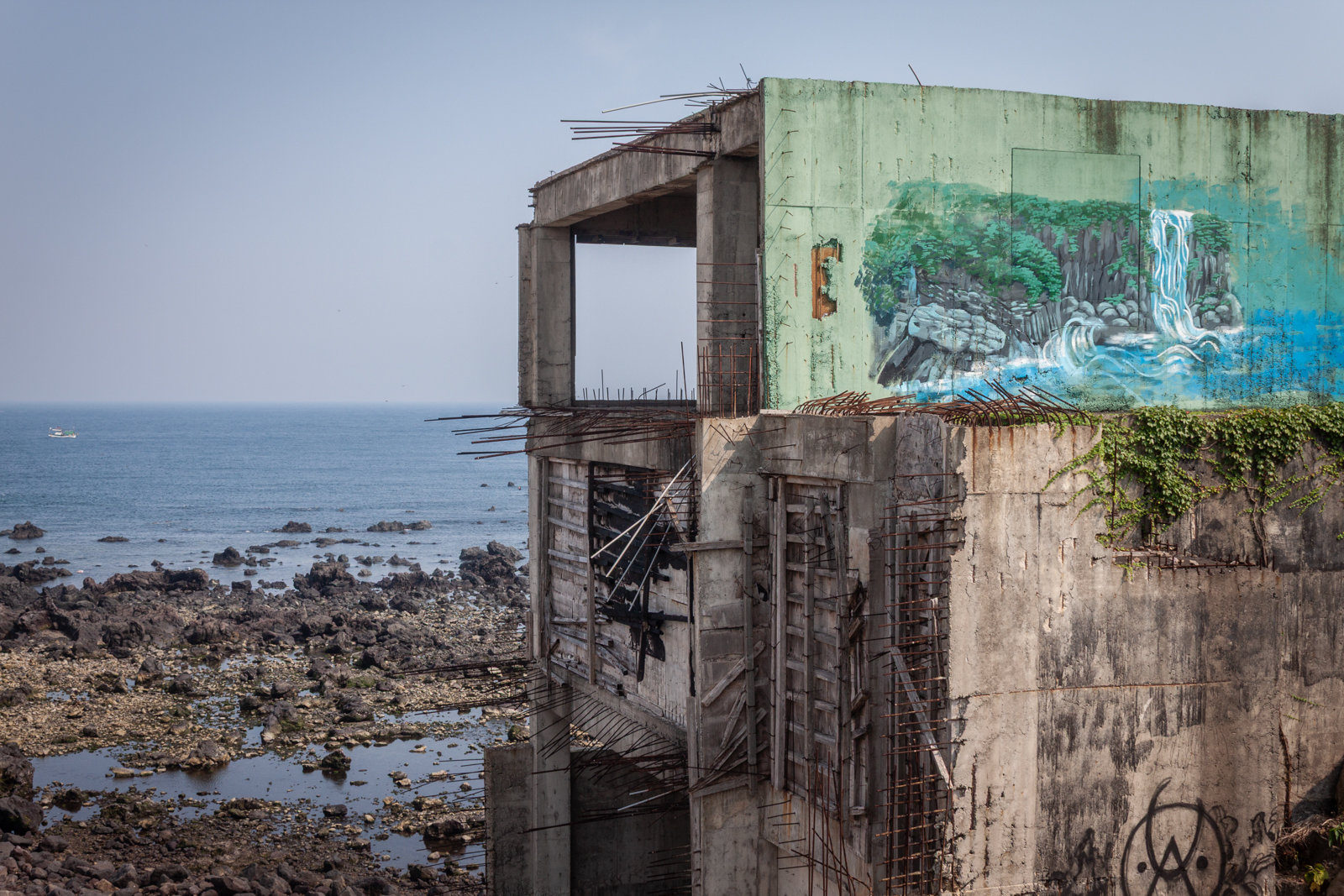 A partially crumbling building with a colorful mural of a waterfall, standing on the rocky shore of the sea.