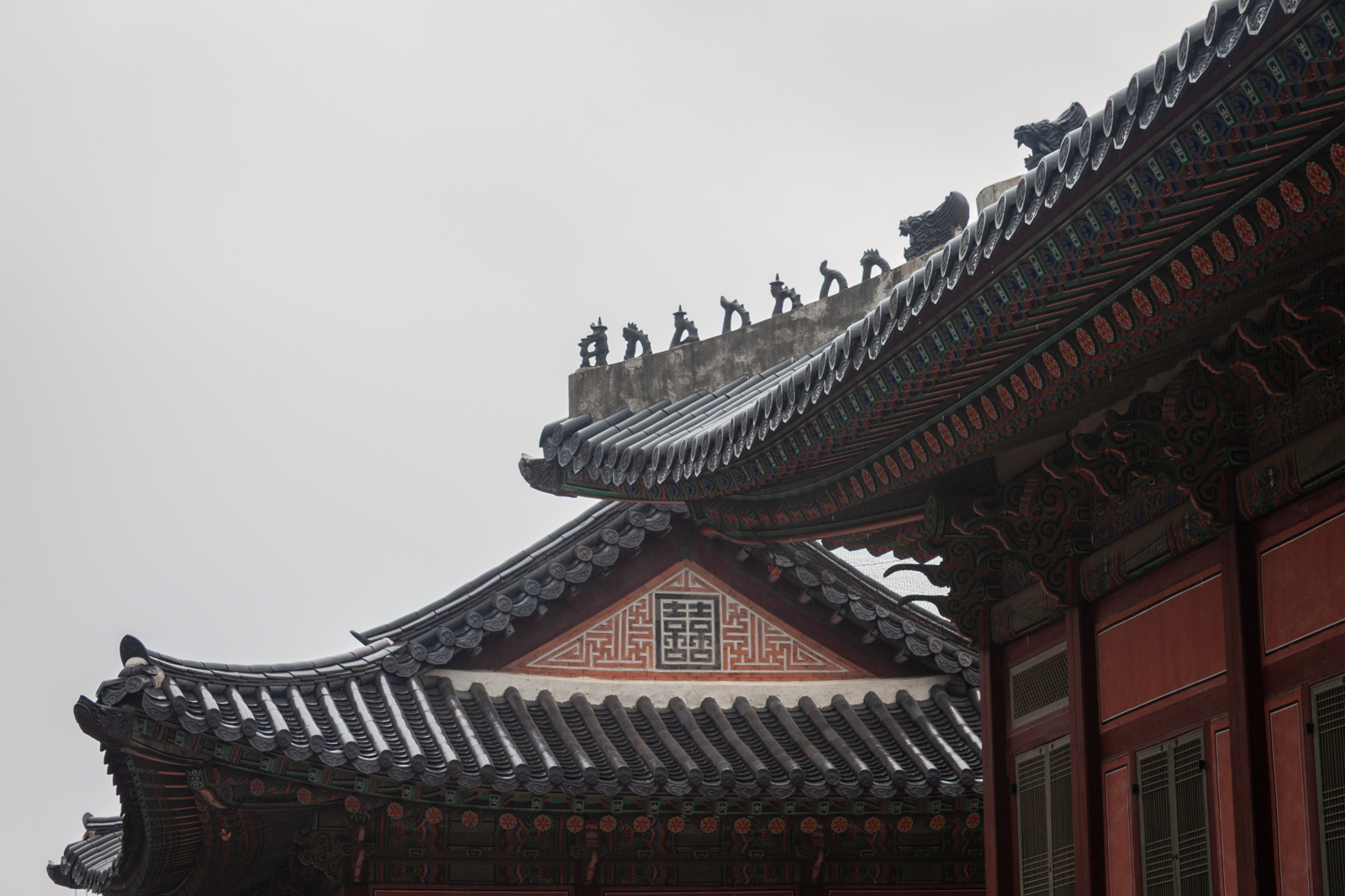 Close-up of ornate rooftop details on a traditional Korean palace. Small statues of animals and mythical creatures line the ridge of the roof, with intricate designs on the eaves below, set against a cloudy sky.
