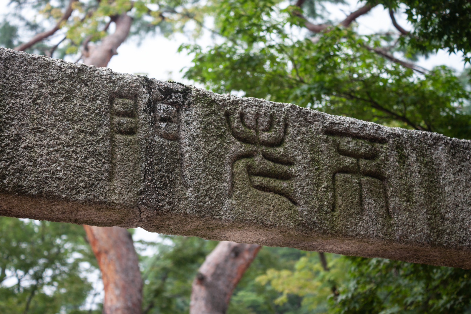 An old stone beam with East Asian characters carved into its surface. The beam is part of an outdoor structure, with trees visible in the background, adding a historic and natural feel to the image.