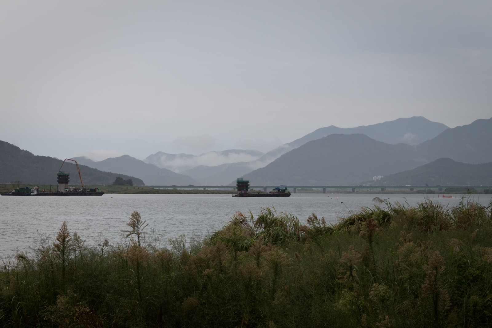 A wide river with mountains in the distance, covered in mist. Two large barges are visible on the water, with a bridge extending across the river in the background, creating a peaceful, natural scene.