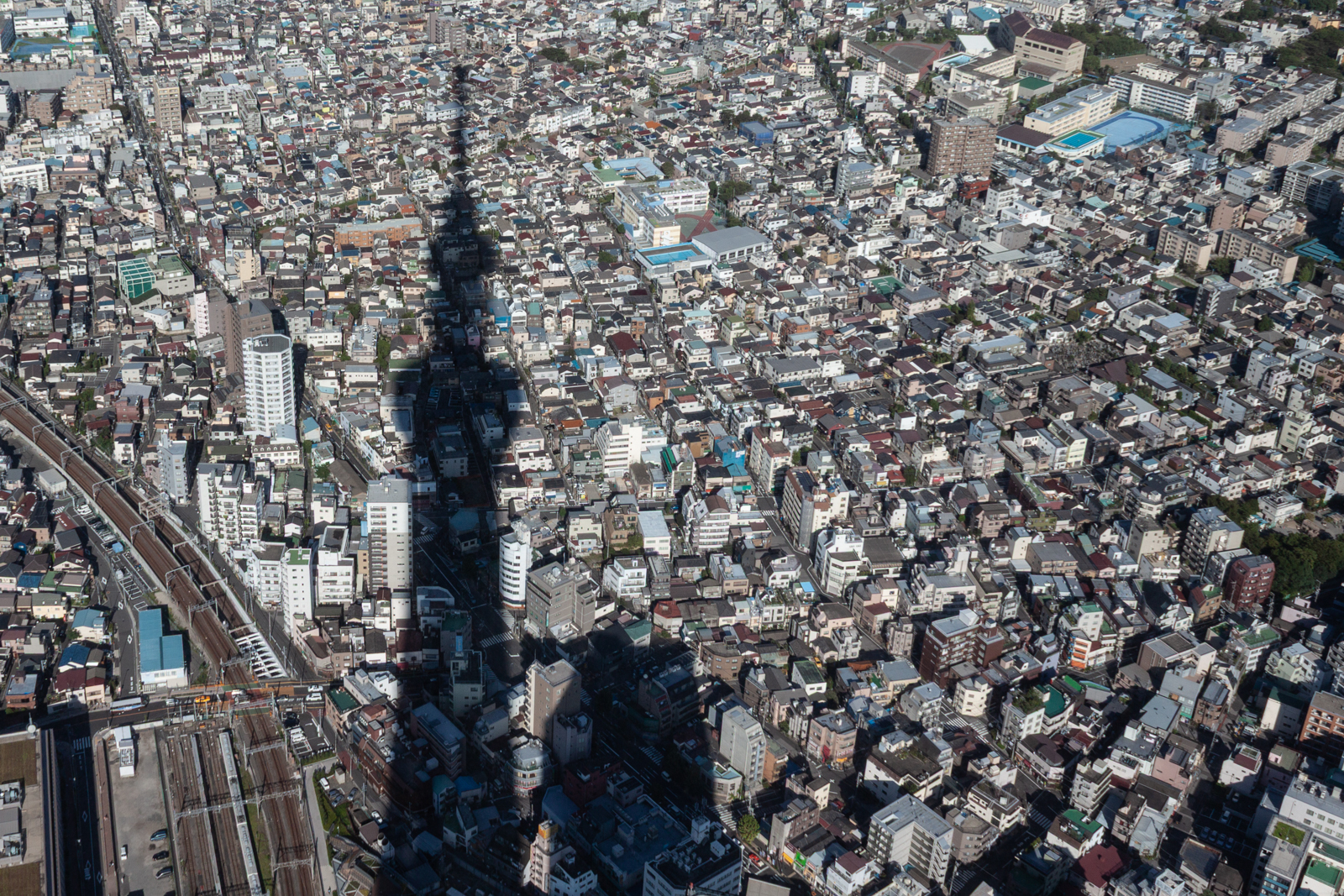 Aerial view of a densely packed urban city with buildings of various sizes. The shadow of a tall structure, possibly a tower, stretches across the cityscape, creating a dramatic contrast against the clustered buildings below.
