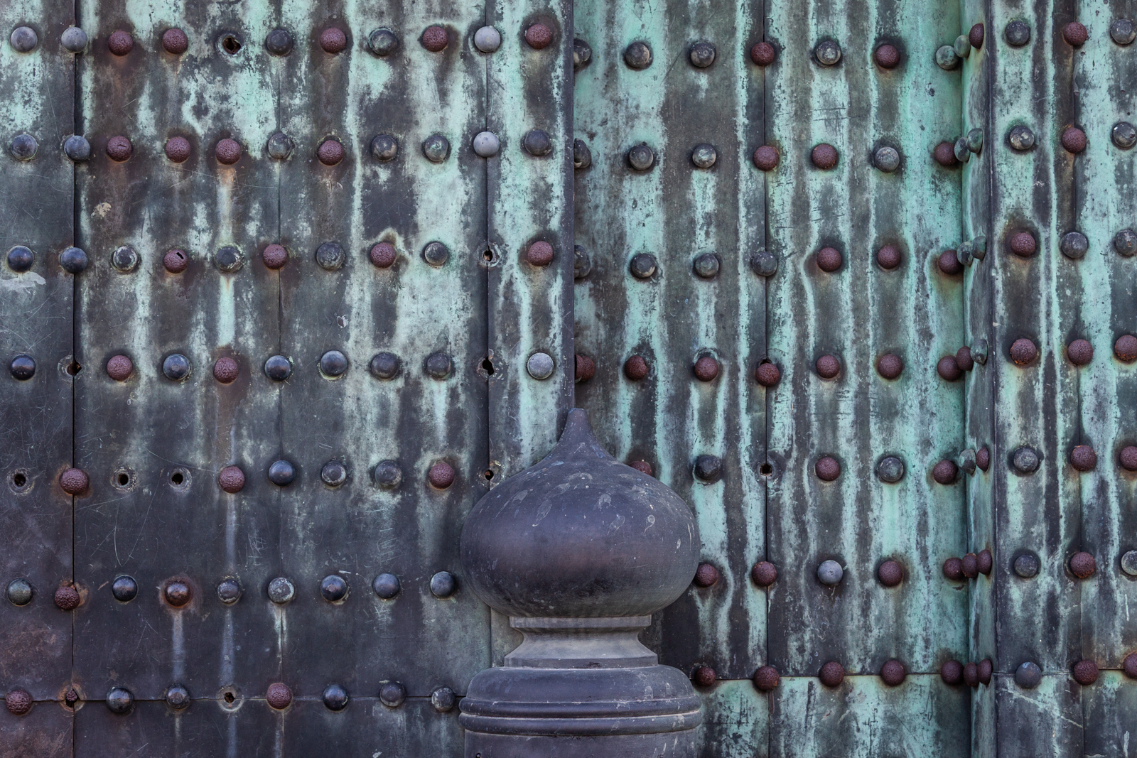 A detailed view of an old, patinated metal gate with rows of rusty rivets. The weathered texture of the gate is highlighted by shades of green and brown, and a decorative stone post stands in front, adding to the historic feel.