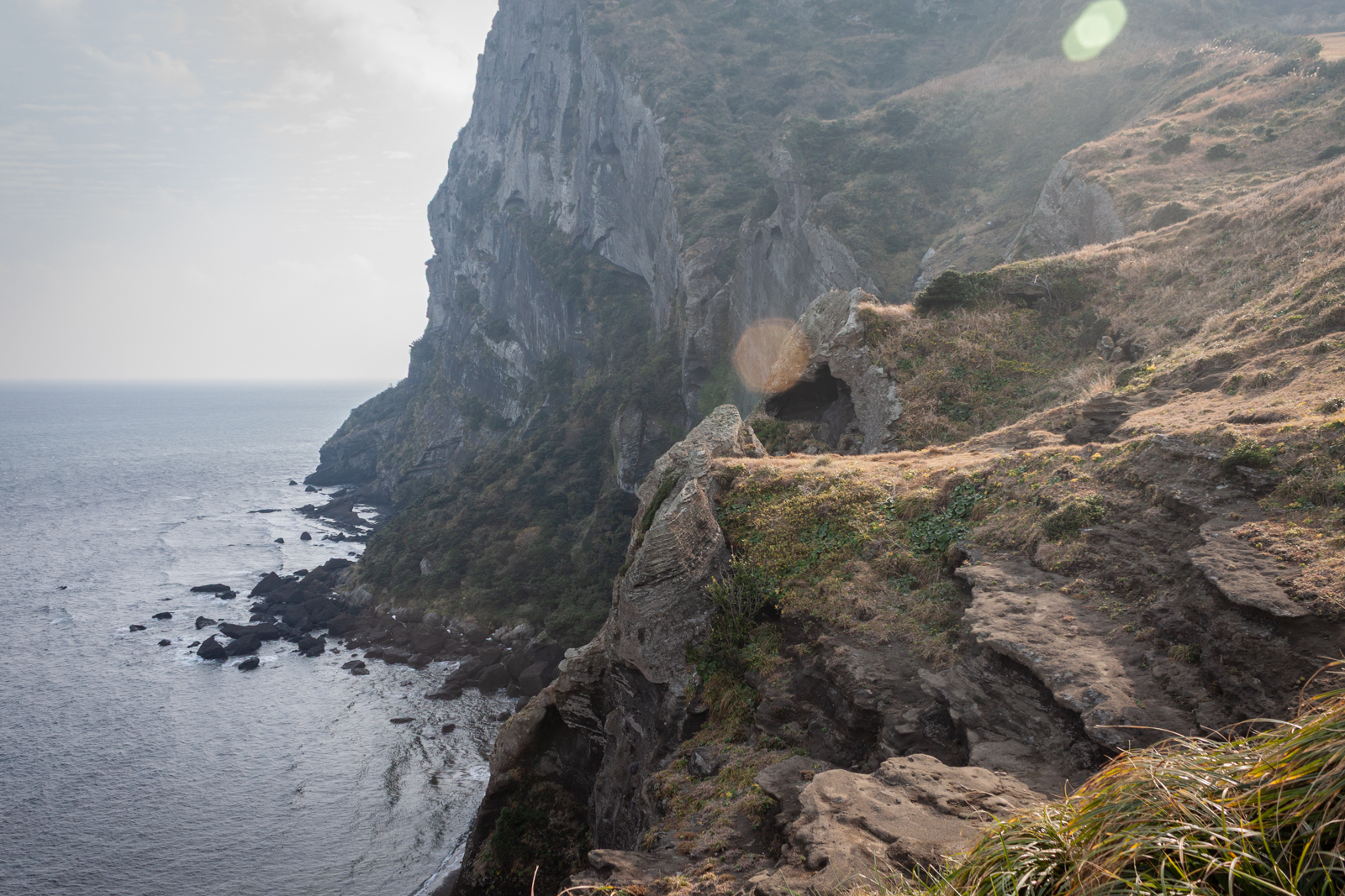 A dramatic coastal cliffside with a rugged path leading up the steep incline. The cliff drops off into the ocean below, where waves crash against the rocks, while patches of sunlight filter through the cloudy sky, casting a soft glow on the landscape.