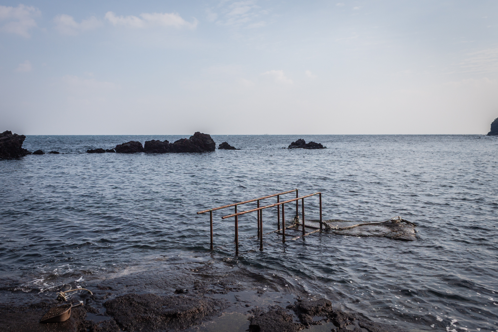 A rocky seashore with an old, rusted handrail extending into the shallow water. The calm sea stretches out toward distant rocks, while the overcast sky creates a serene, contemplative atmosphere.