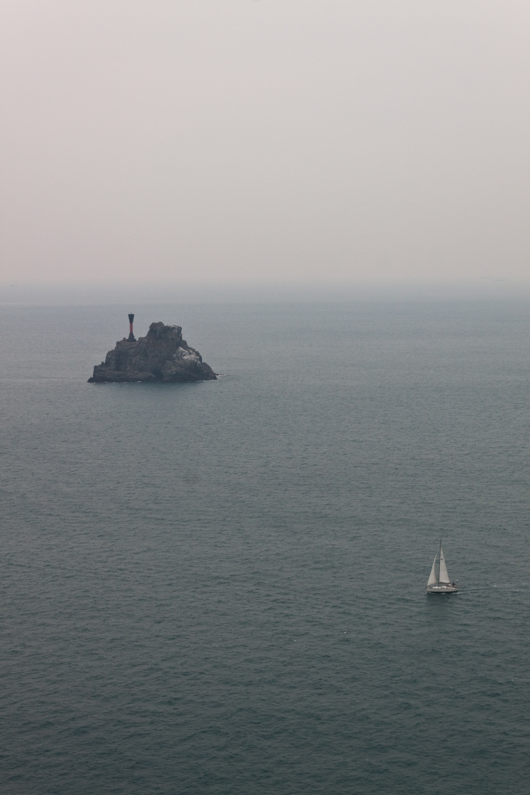 A lone sailboat navigating across a calm, expansive ocean, with a small rocky island featuring a prominent beacon in the distance. The sky appears overcast, casting a muted tone over the scene.