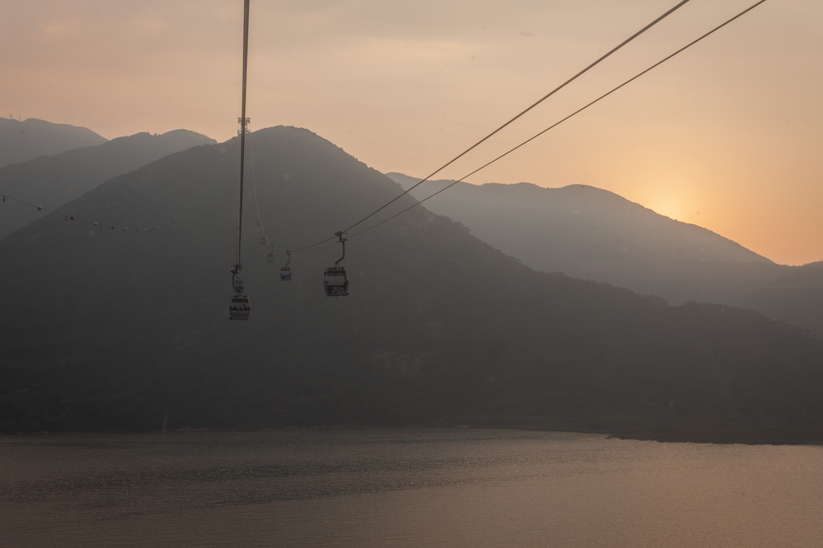 Cable cars gliding over a large body of water at sunset, with silhouetted mountains in the background. The fading sunlight creates a soft, golden hue across the sky and water.