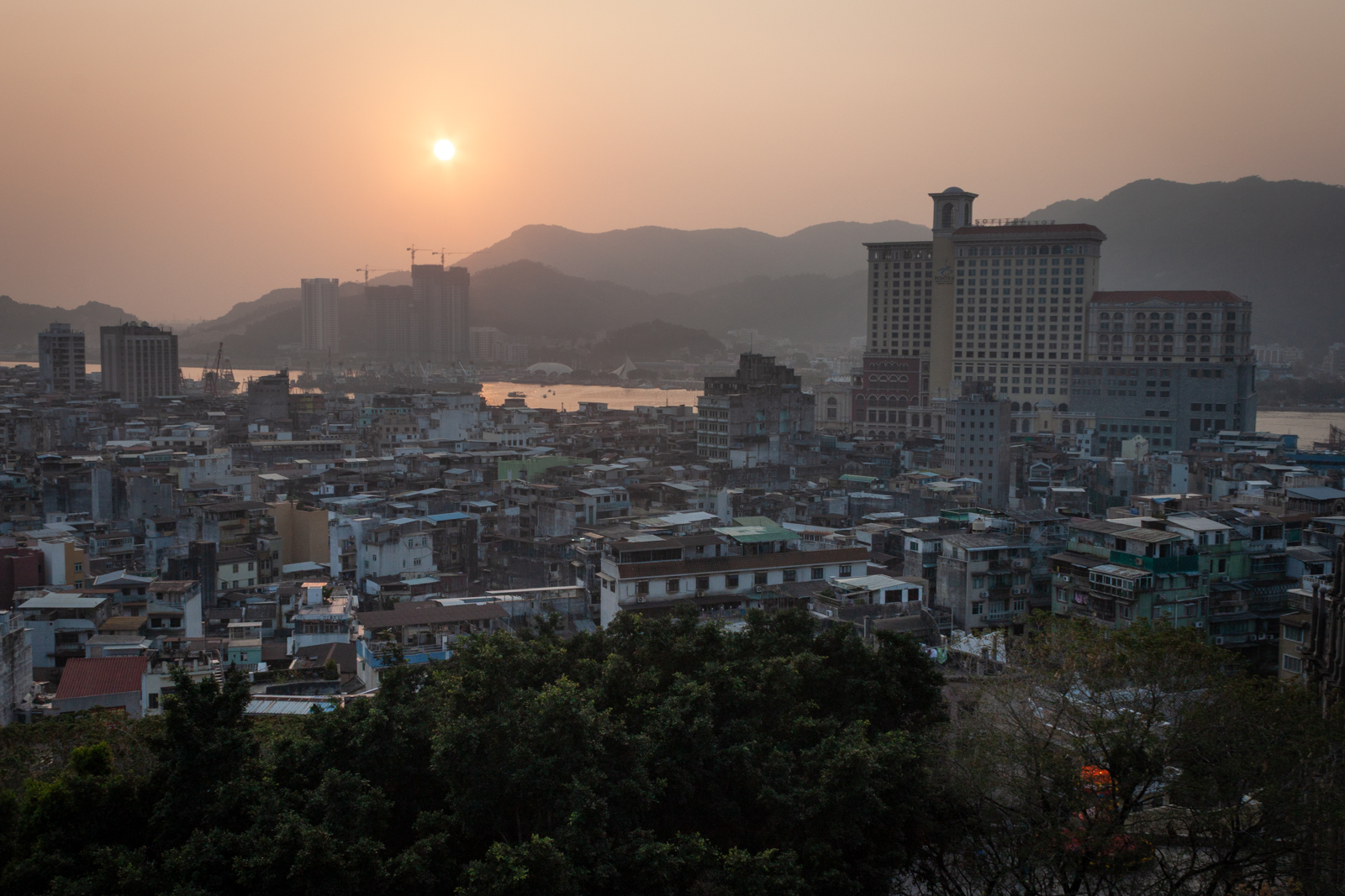 A hazy sunset over a densely packed urban landscape, with low-rise buildings in the foreground and taller structures in the distance. Mountains and a river can be seen beyond the city, softly lit by the setting sun.