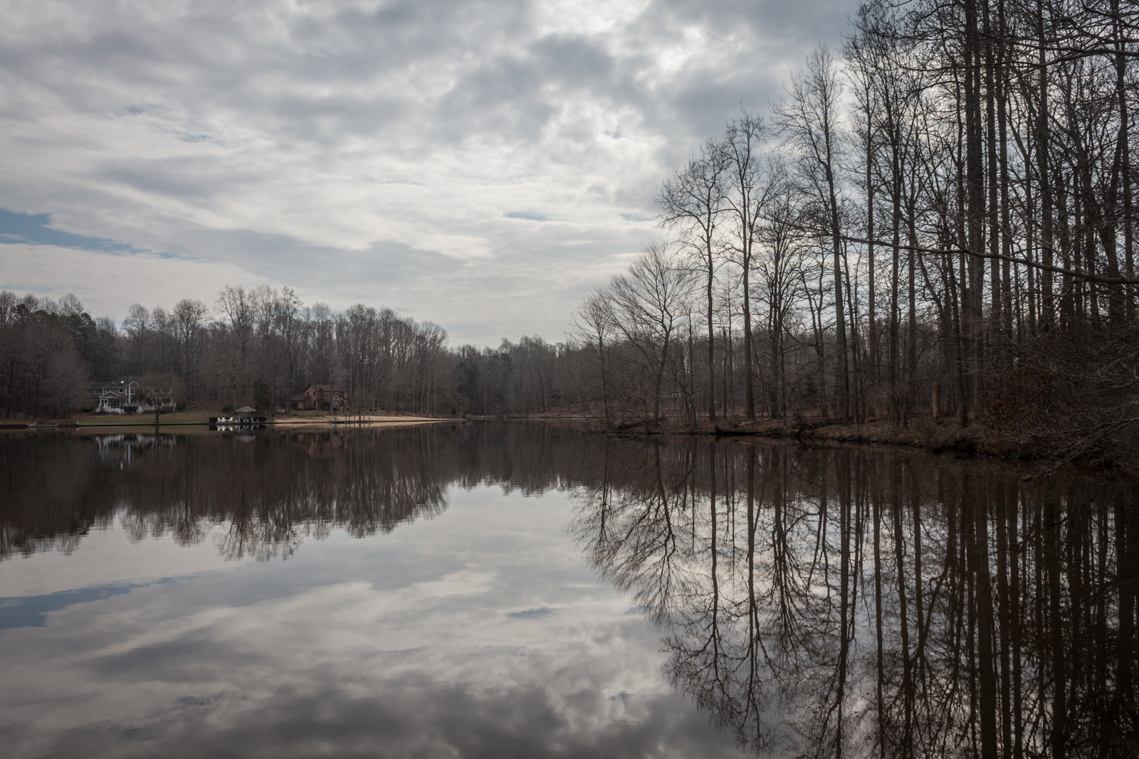 A calm lake reflects the overcast sky and the trees lining the shore. The still water mirrors the leafless trees, while houses and docks sit quietly along the far side of the lake, adding to the peaceful winter landscape.