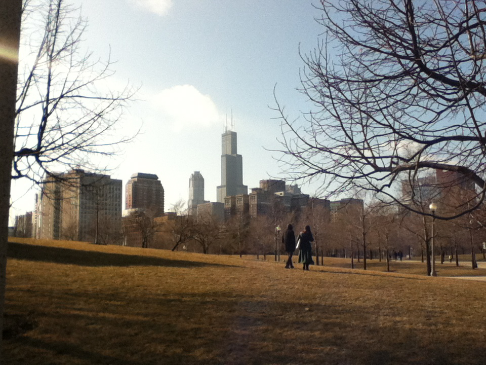 A color photo taken from a park, showing the Chicago skyline in the distance. Two people are walking along a path lined with bare trees. The sun shines brightly, casting long shadows on the grassy foreground.