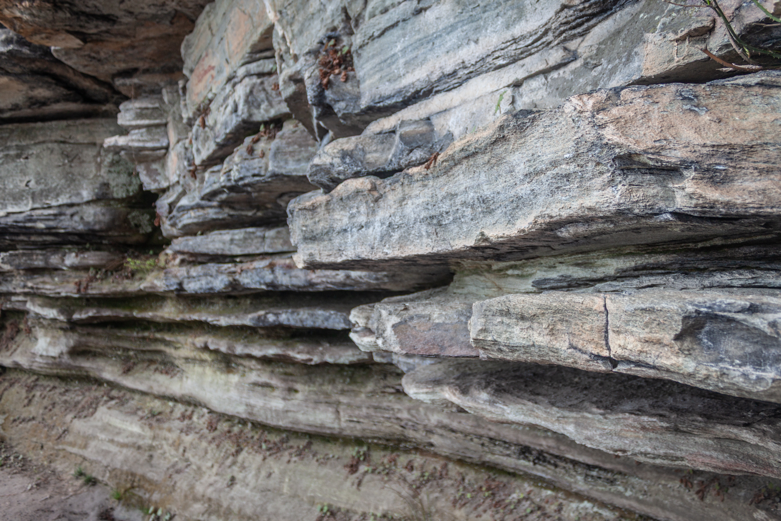 A close-up view of stratified rock layers with varying shades of gray and brown, showing signs of erosion and weathering. The flat, horizontal layers are stacked, creating a rugged, textured appearance.