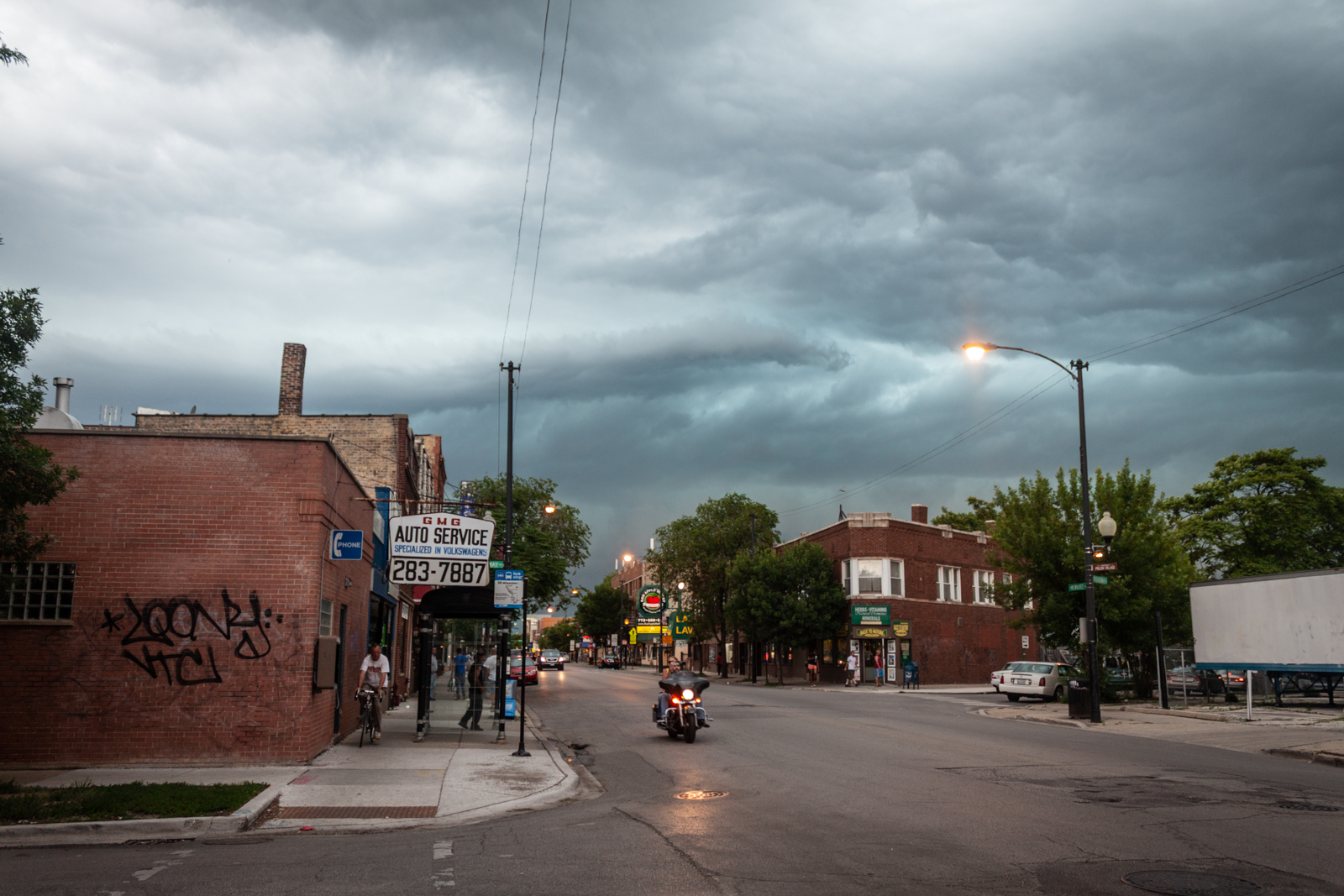 A wide-angle view of a quiet urban street under a dark, stormy sky. The street is lined with brick buildings, including an auto service shop with a prominent sign and graffiti on its side. A motorcyclist is seen riding down the road, while pedestrians walk on the sidewalk, giving a sense of calm before the impending storm. The ominous clouds contrast with the soft streetlights, creating a dramatic atmosphere.