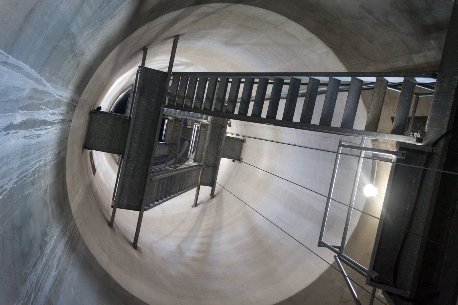 A steep, industrial staircase spirals upwards inside a large, cylindrical concrete shaft. The metal stairway is sharply angled, creating a dramatic perspective that draws the eye toward the distant opening at the top. The walls of the shaft are smooth, and faint streaks of water stains run along the surface, adding texture to the otherwise stark surroundings. The lighting is minimal, with a single bare bulb casting shadows and emphasizing the utilitarian design of the structure.