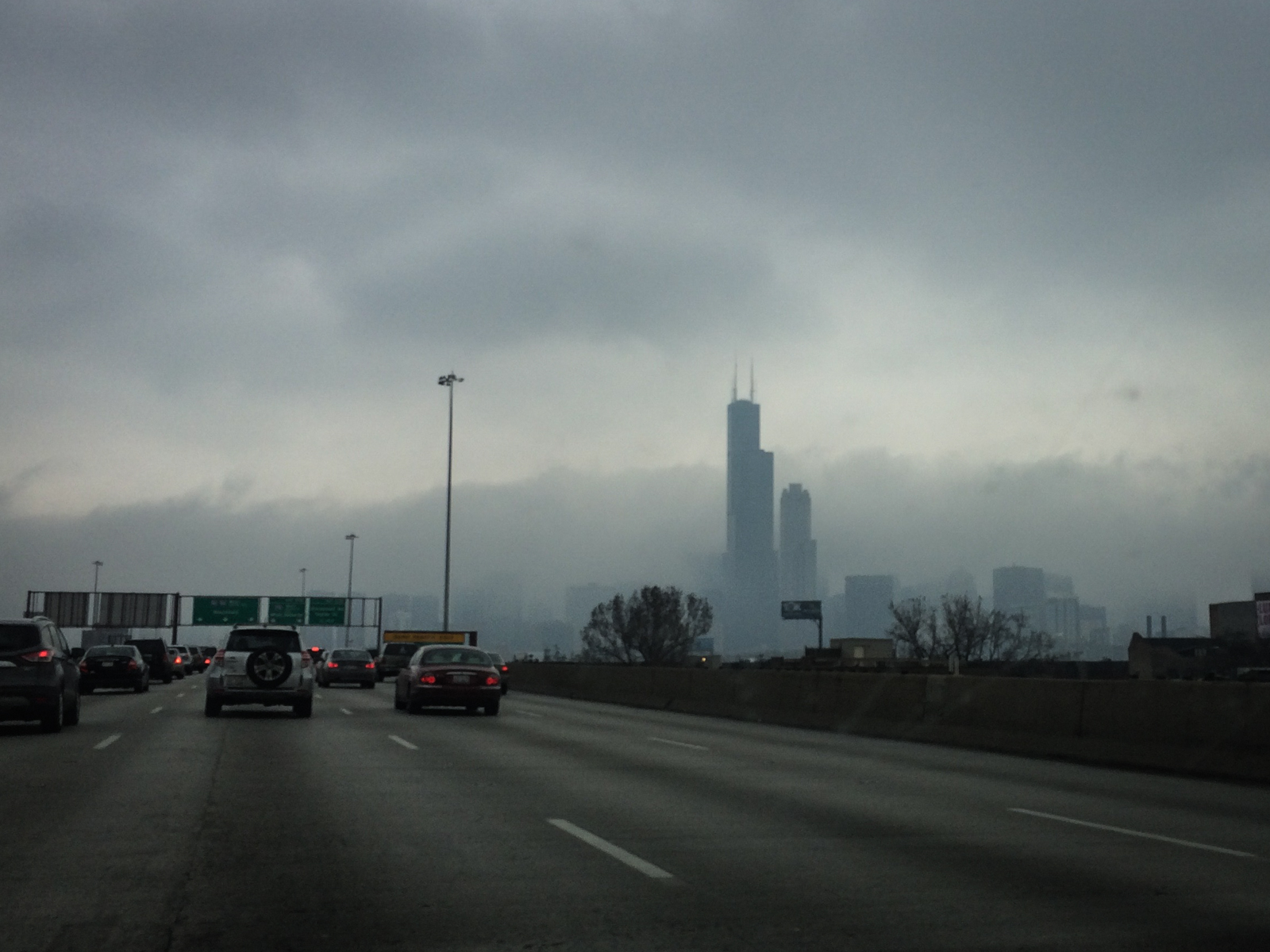 View from a car on a highway heading toward a fog-covered city skyline, with the silhouette of a tall skyscraper, the Willis Tower, faintly visible through the mist. The sky is overcast, and the road is lined with other vehicles.