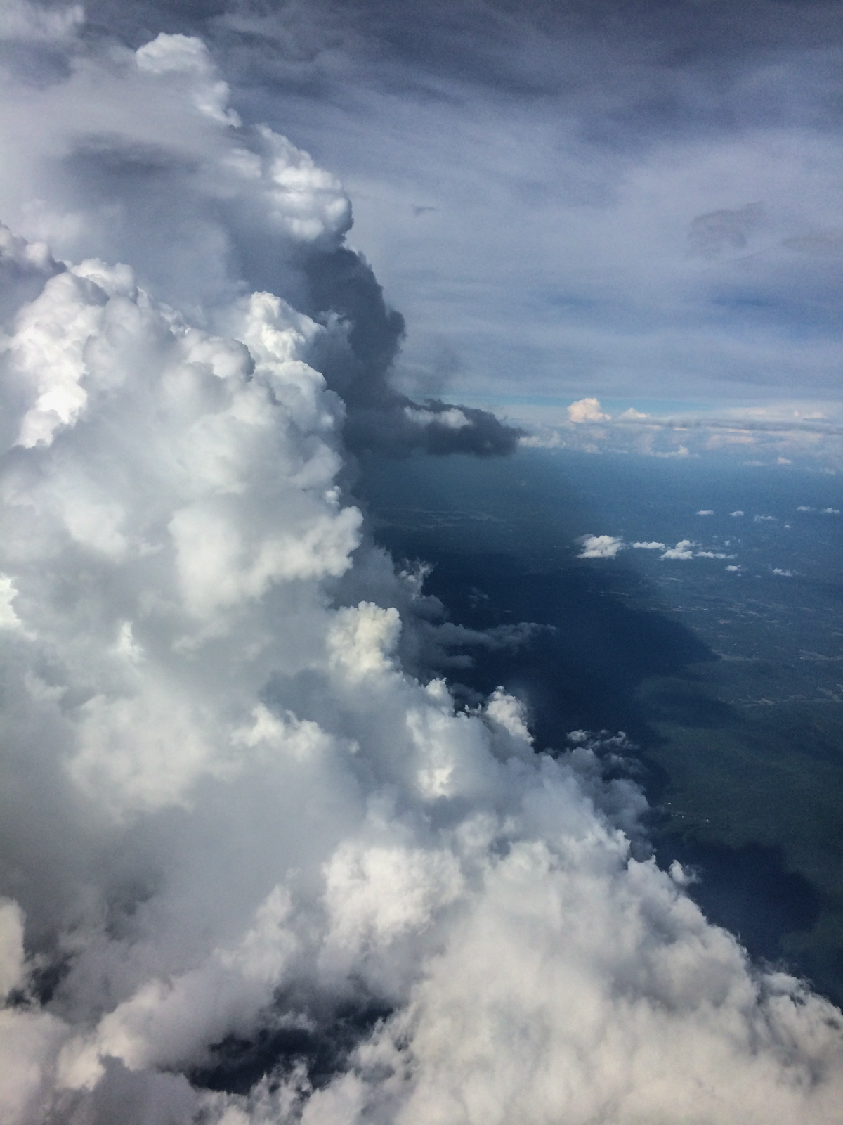 View of large, billowing clouds from above, taken from an airplane. The towering clouds cast shadows over the landscape below, with a bright blue sky in the distance.