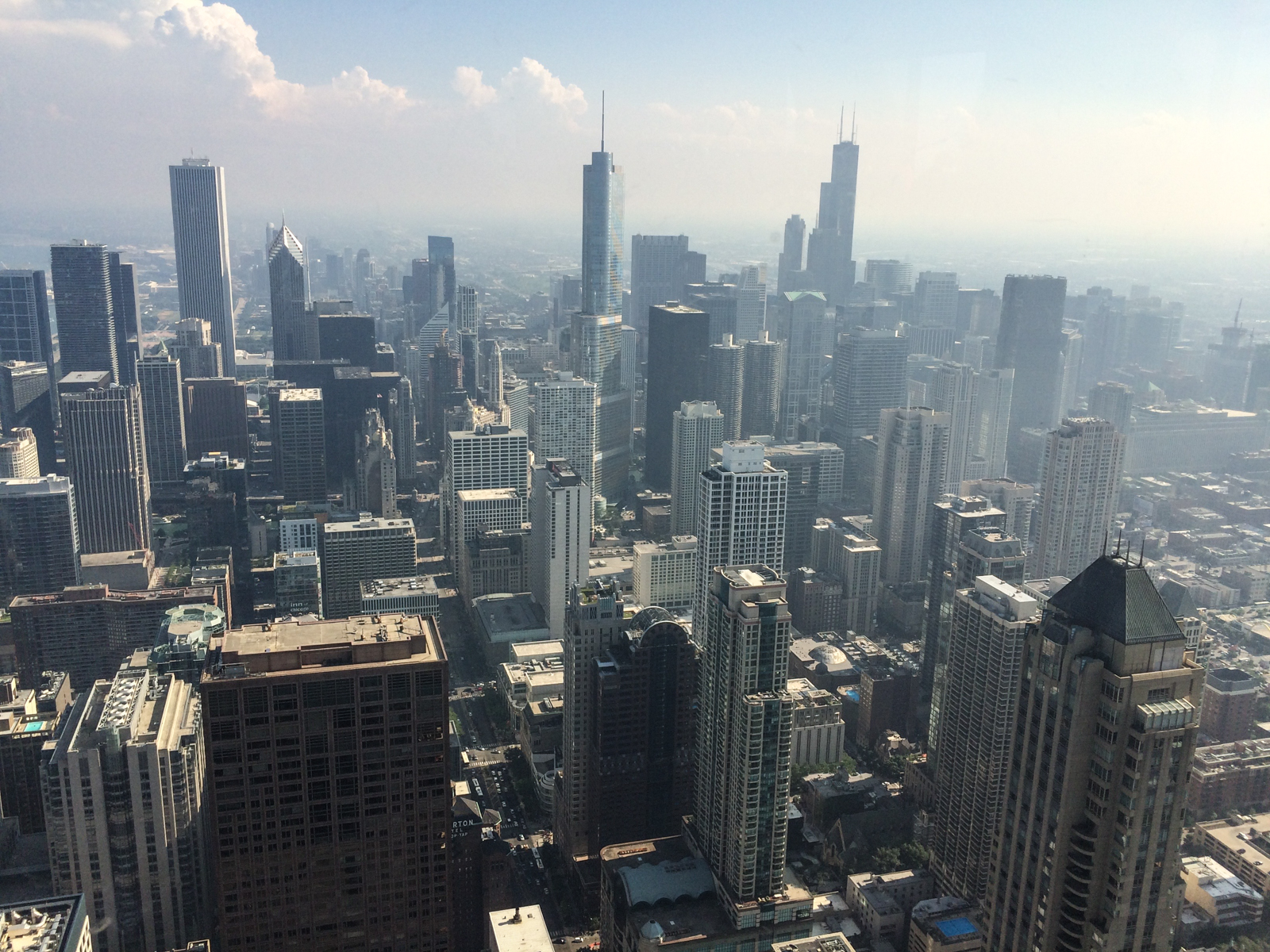 Aerial view of a bustling city skyline on a clear day, showcasing numerous skyscrapers and high-rise buildings, with the Willis Tower standing tall in the background. The sky is slightly hazy, with soft clouds forming in the distance.