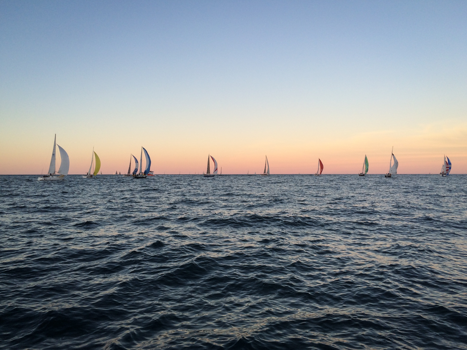 Several sailboats with colorful sails scattered across the water at sunset. The sky is a gradient of soft pinks, oranges, and blues, while gentle waves ripple across the surface of the sea.