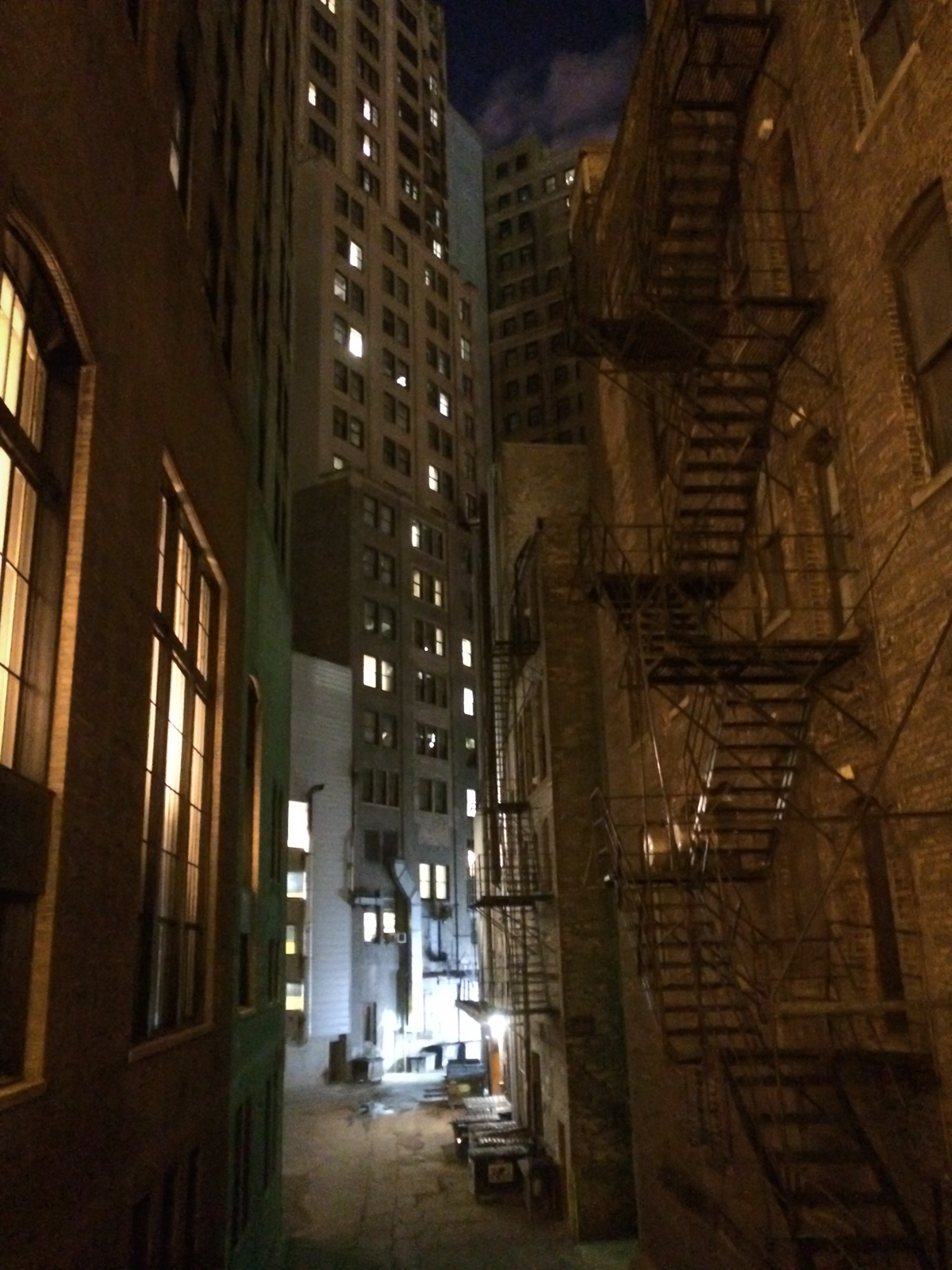 Dimly lit urban alleyway at night, surrounded by tall buildings with fire escapes on one side and windows glowing with light. The alley is empty, with trash bins near the far end, and the tops of skyscrapers rise into the night sky.