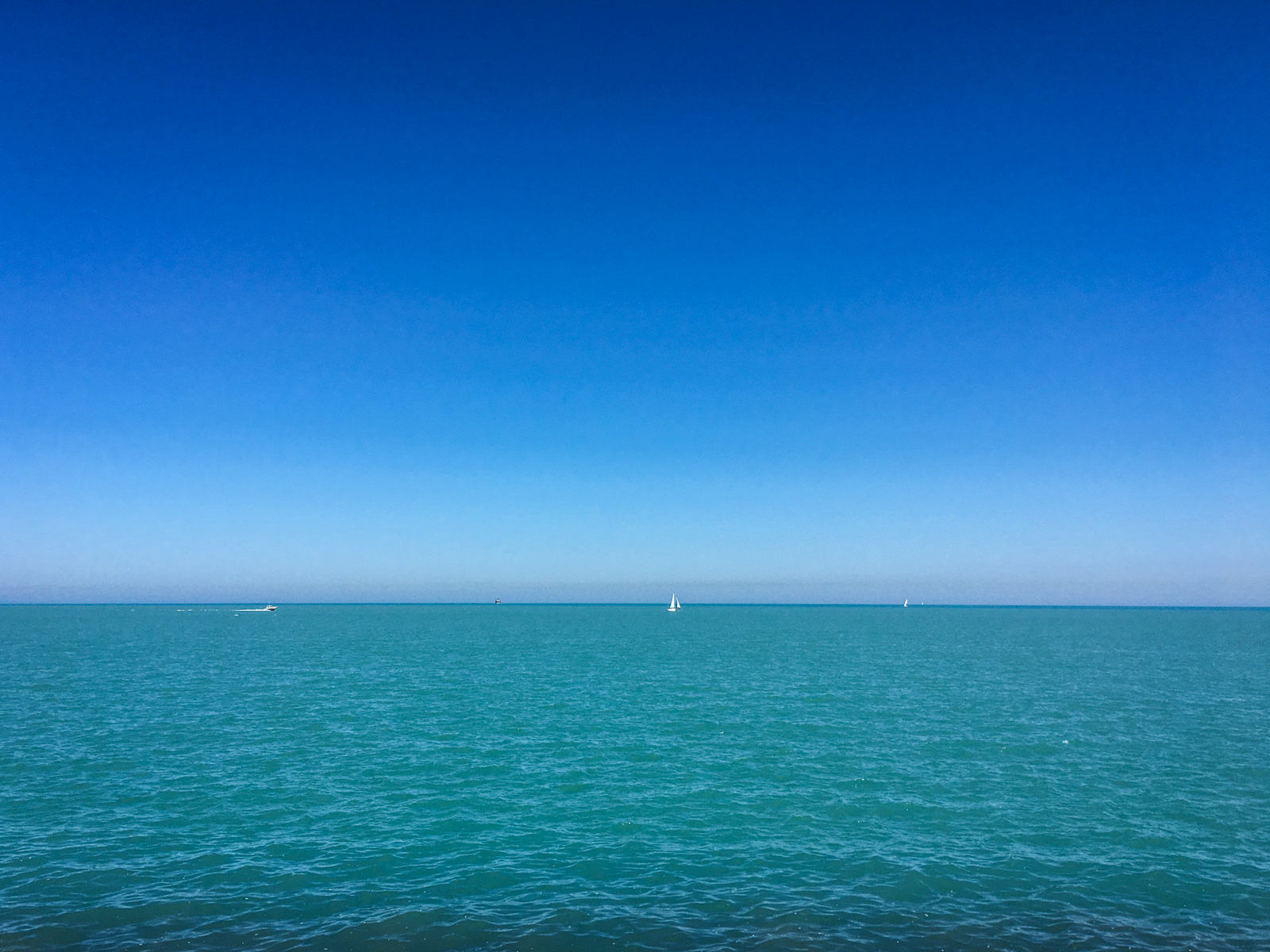 Clear, bright day over a calm turquoise sea, with a deep blue sky stretching above. A few small sailboats and distant vessels can be seen on the horizon, creating a peaceful and serene scene.