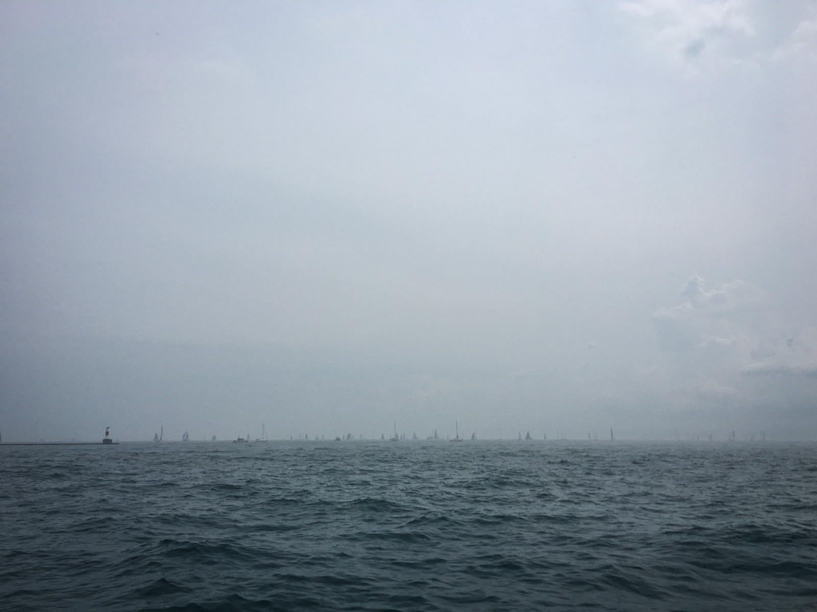 A hazy day over a calm ocean, with distant sailboats barely visible on the horizon and a faint lighthouse to the left, blending into the foggy atmosphere.