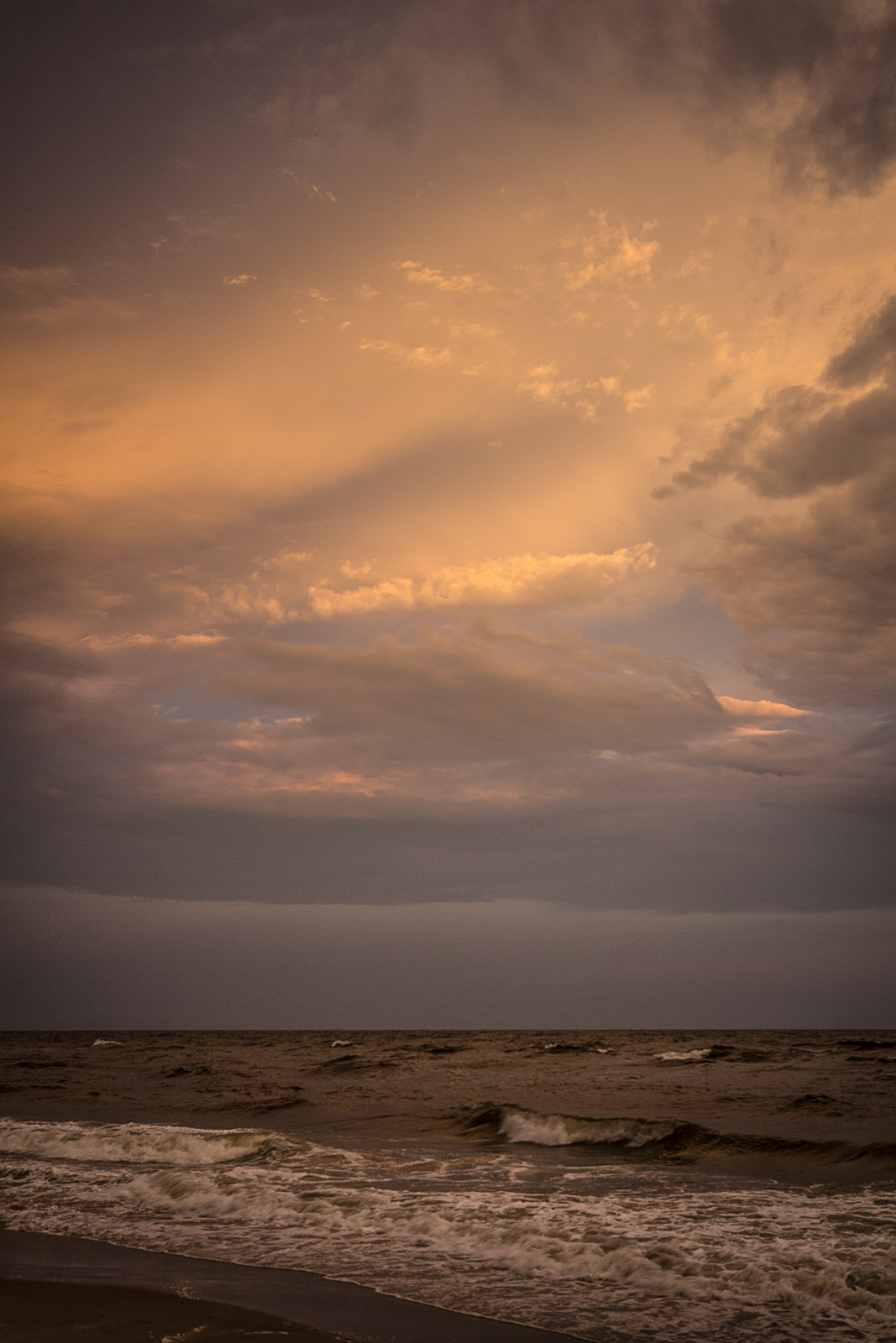Dramatic seascape at dusk with waves crashing on the shore and a sky filled with layered, glowing clouds in shades of orange, gold, and purple.