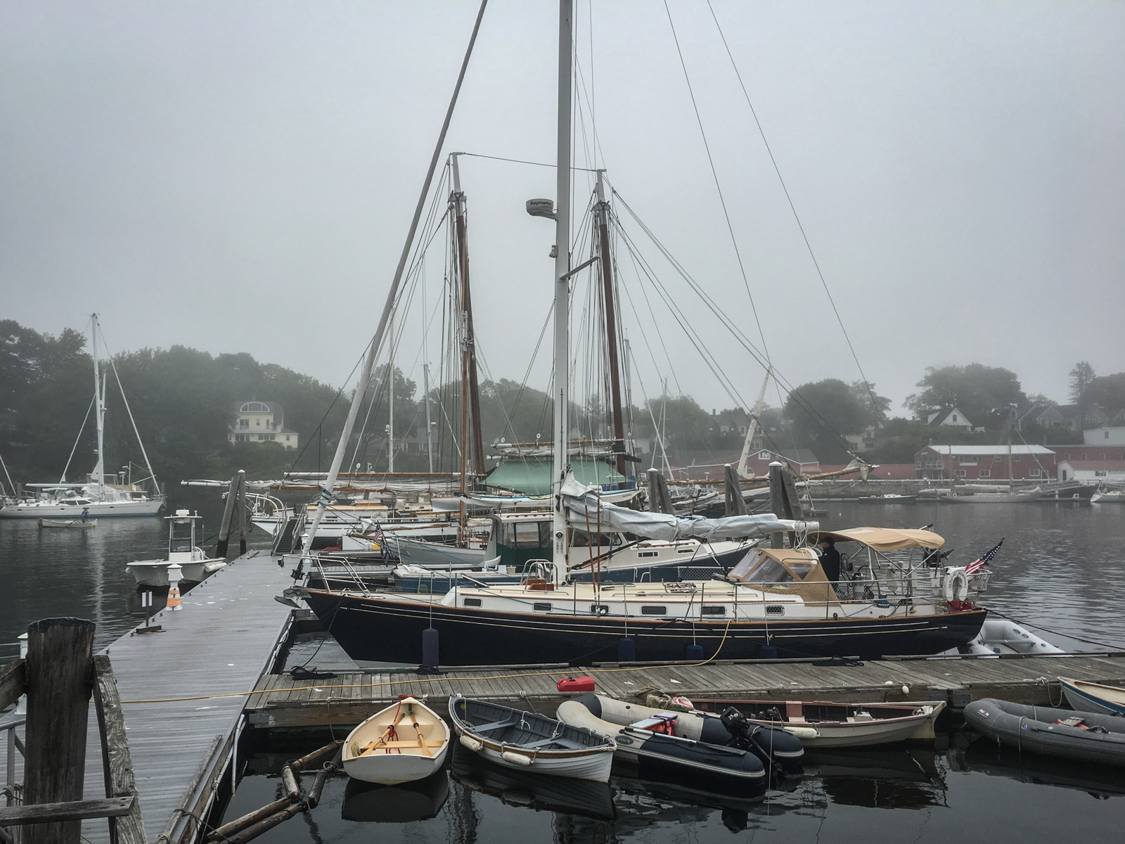 A foggy marina scene with several sailboats docked along a pier. Smaller boats are tied up in the foreground, while the distant shoreline and houses are barely visible through the mist. The atmosphere is quiet and calm, with muted tones.