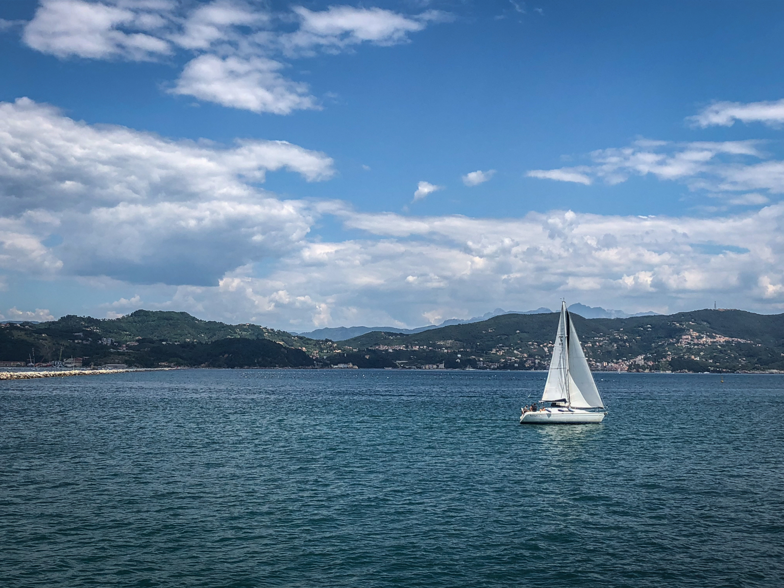A lone sailboat gliding across calm blue waters, with a picturesque mountainous coastline in the background.
