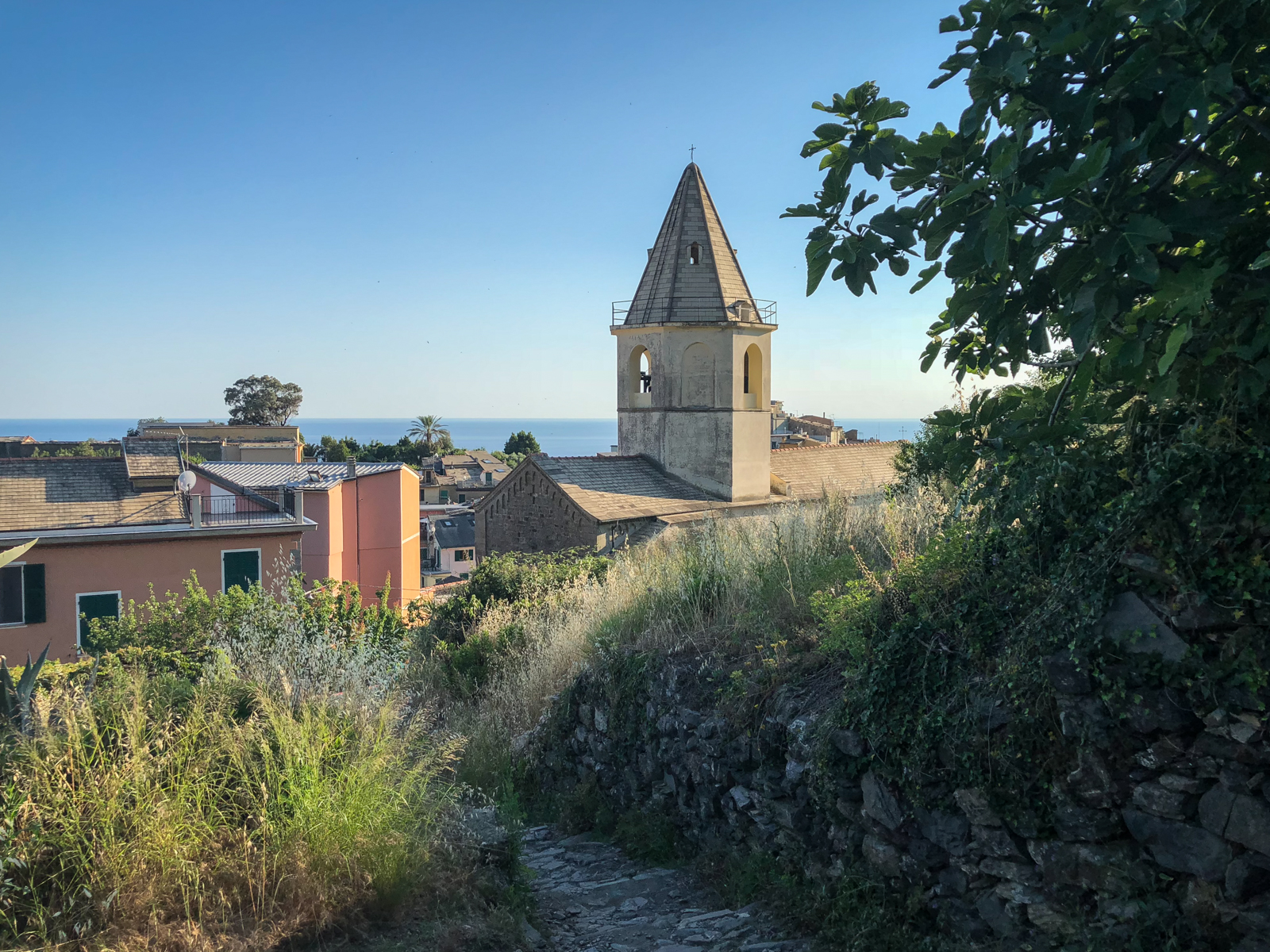 A charming church tower in a small village with the blue sea visible in the background. The image has a calm, picturesque feeling, with rustic houses lining the narrow pathways of the village.