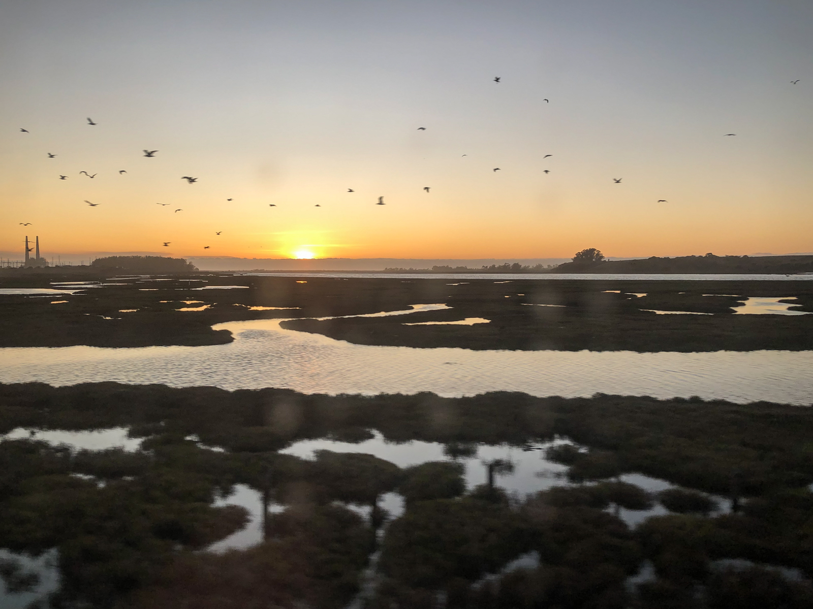 A tranquil sunset over a marshland with patches of water reflecting the soft light. The sky is a gradient of orange and yellow as the sun dips below the horizon. A flock of birds is in flight across the sky, adding movement to the peaceful scene. In the distance, silhouettes of trees and industrial structures, including a pair of tall chimneys, can be seen, contrasting with the natural landscape. The calm waters and open space evoke a serene and quiet atmosphere.