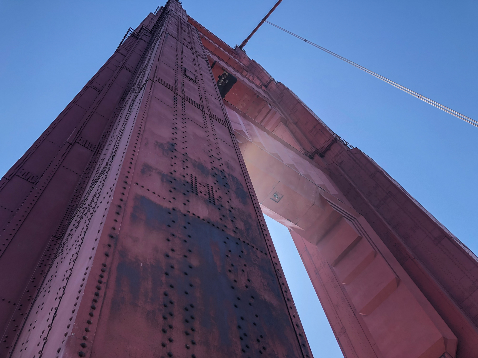A close-up, upward view of a massive red tower of a suspension bridge. The riveted steel beams and weathered surface of the tower are bathed in sunlight, with cables extending from the structure into the clear blue sky above. The sheer size and industrial details of the tower dominate the image.