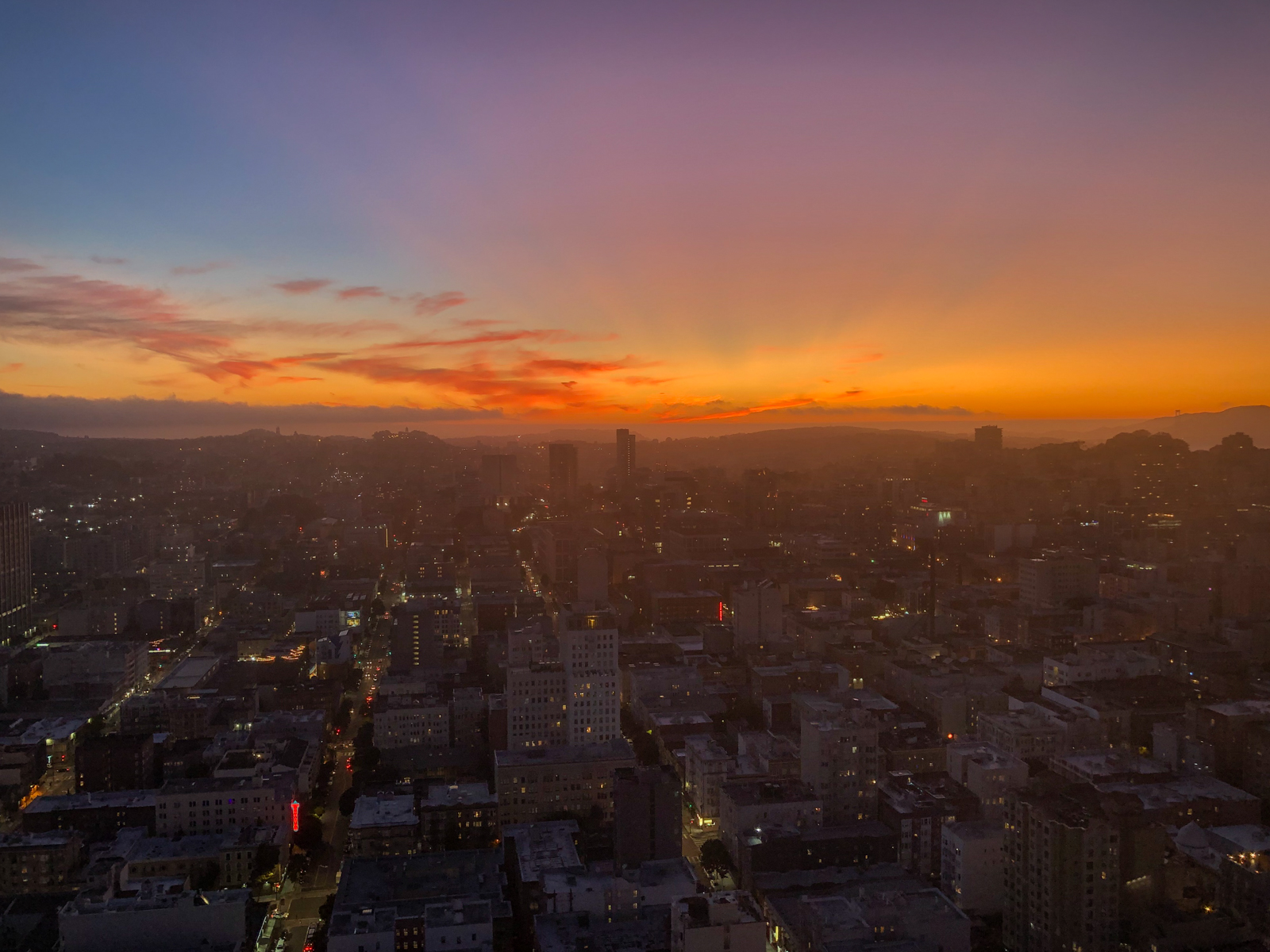 A breathtaking cityscape at sunset, with the sky awash in vibrant shades of orange, pink, and purple as the sun dips below the horizon. The city below is beginning to light up for the evening, with illuminated streets and buildings visible as the twilight deepens. In the distance, hills and a faint outline of water are visible, with the soft glow of the sunset casting a warm hue over the entire scene.