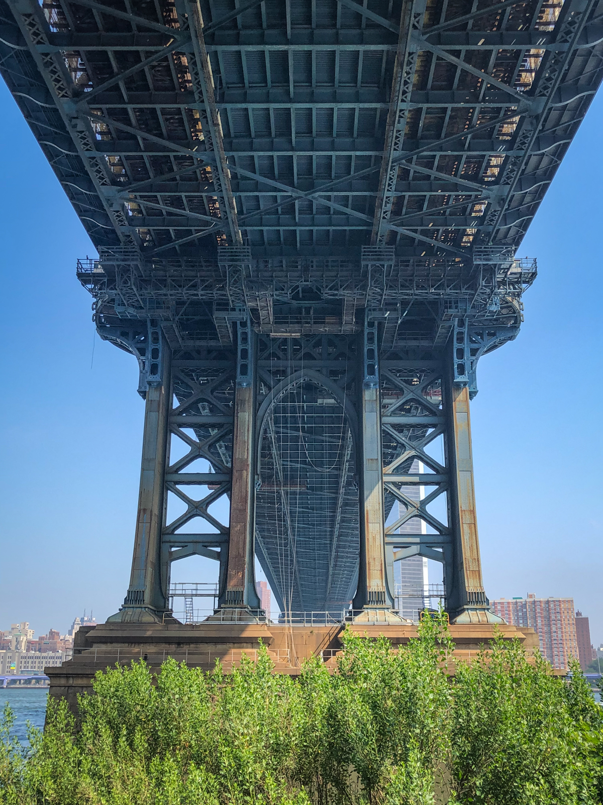 A dramatic view from beneath a large suspension bridge, showcasing the intricate metal framework and beams that support the structure. The massive pillars of the bridge rise from a base surrounded by greenery, with the city skyline faintly visible in the background under a clear blue sky. The perspective emphasizes the scale and engineering of the bridge, creating a sense of both strength and complexity.