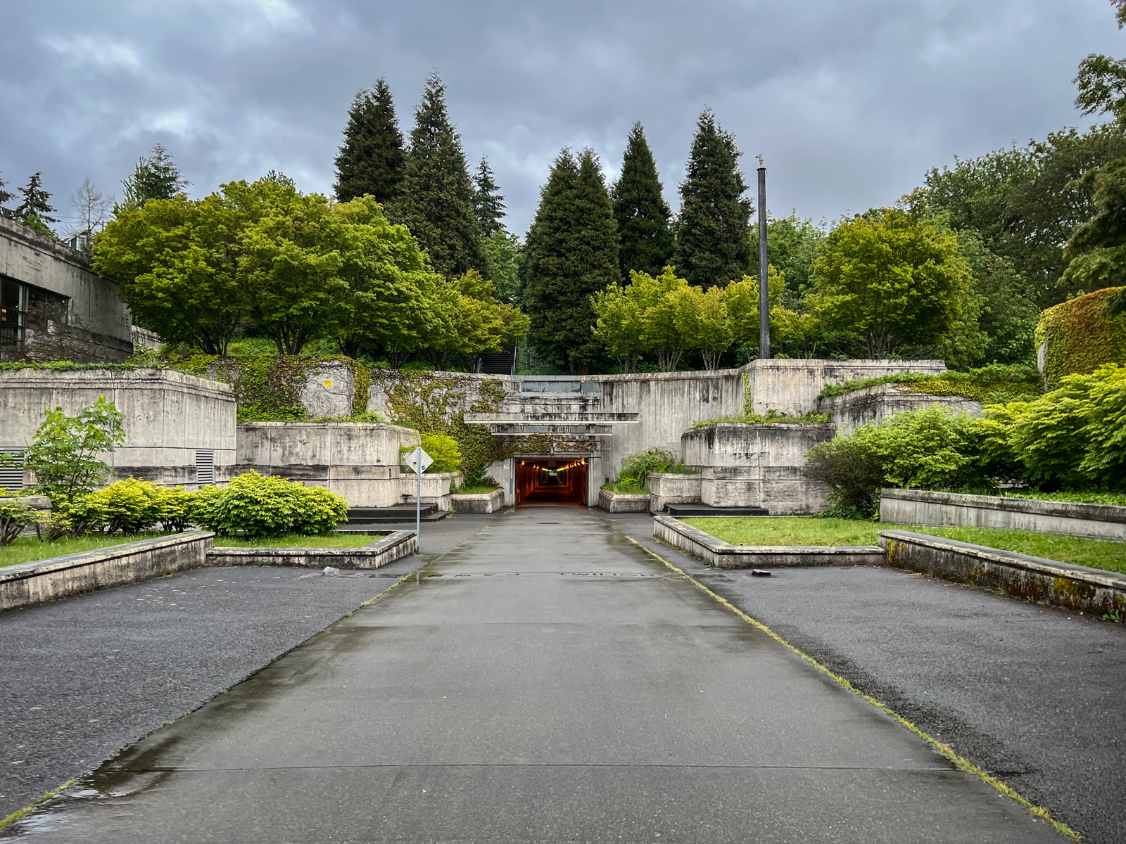 A concrete, terraced landscape with greenery and trees arranged in levels. The pathway leads into a tunnel, flanked by dense foliage and industrial design elements. The scene is slightly overcast, adding a moody, contemplative feel to the structured, urban nature of the setting.