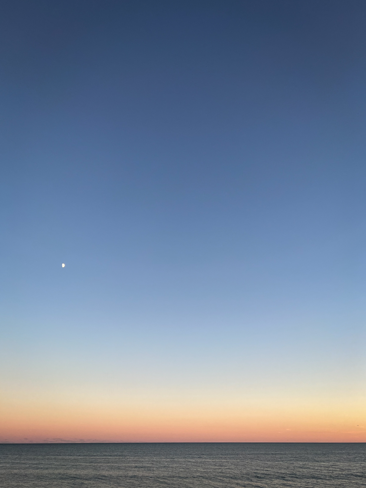 A serene image of a vast ocean horizon at sunset, with the sky transitioning from soft pastel orange near the horizon to deep blue higher up. A small, bright moon is visible in the upper left corner, contrasting with the fading daylight. The calm water reflects the warm hues of the setting sun, creating a peaceful and minimalist scene, emphasizing the tranquility of nature.