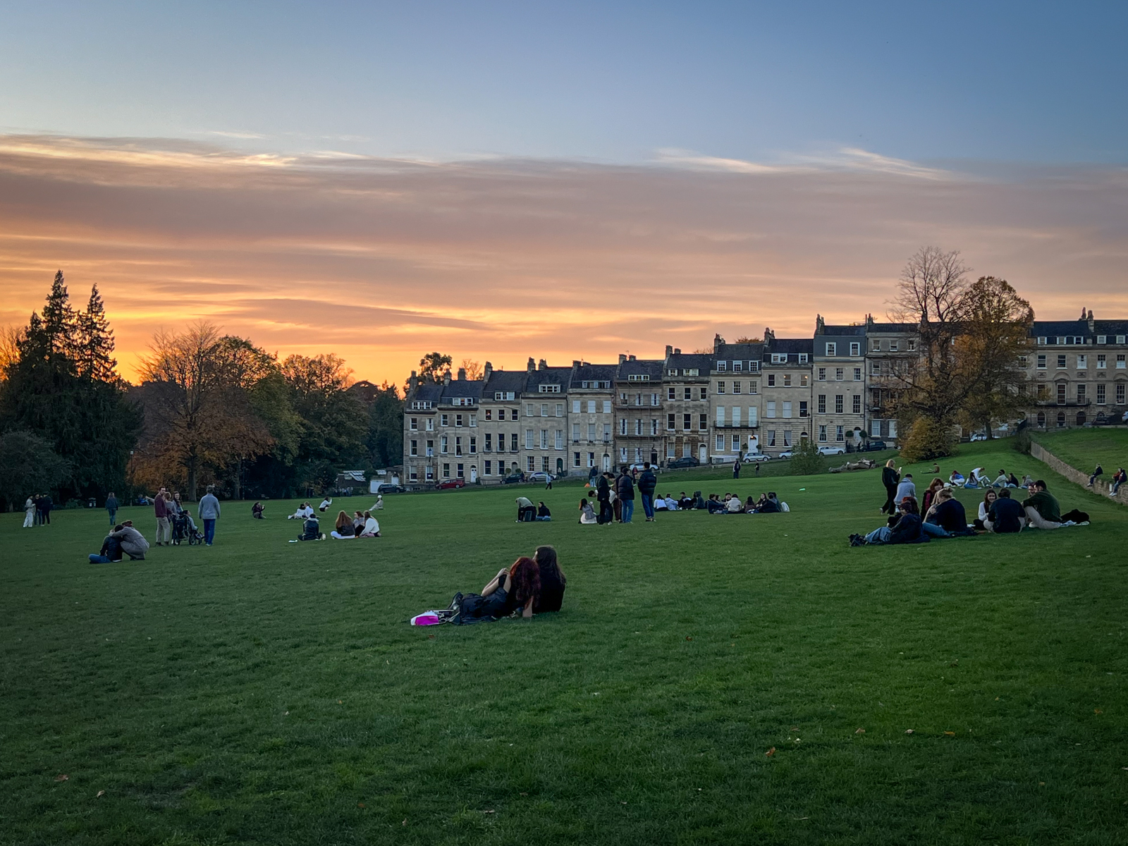 A tranquil sunset scene in a park, with groups of people relaxing on the expansive grassy field. In the background, historic buildings with stone facades line the horizon, and the sky is filled with warm orange and pink hues, transitioning into a soft blue. The trees in the park add an autumnal touch, with some displaying golden leaves as the sun sets.
