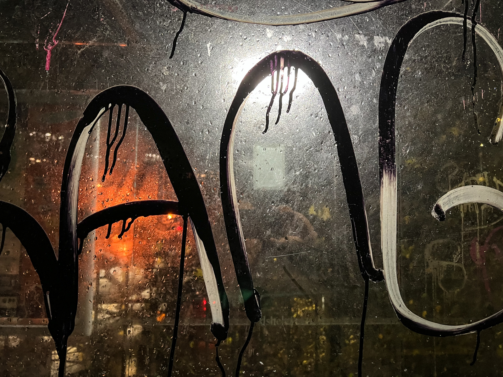 A close-up of a glass window covered in large black graffiti, with light shining through from behind, highlighting the droplets of moisture and creating a wet, gritty texture. An orange light glows faintly in the background.