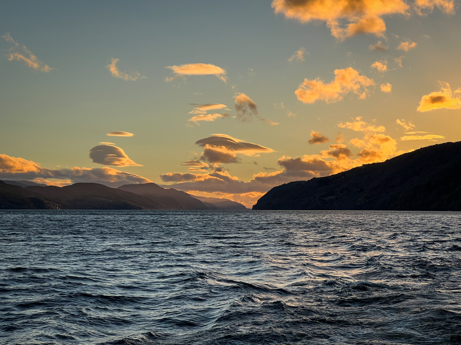 A serene view of a large body of water surrounded by hills at sunset. The sky is filled with scattered clouds illuminated by the warm glow of the setting sun, casting soft reflections on the water's surface.